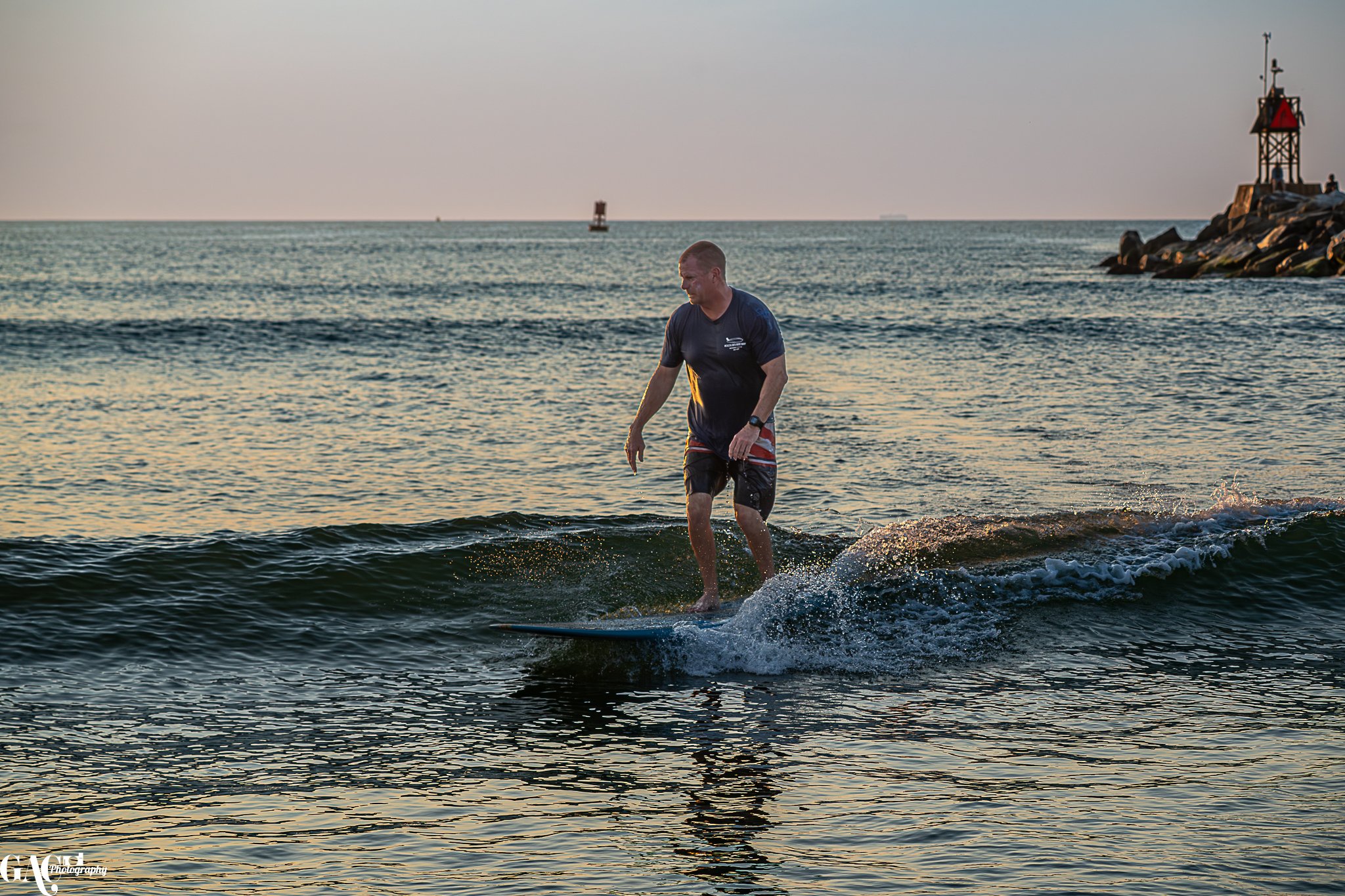 A man surfing on a wave near the shoreline during sunset, with a lighthouse and rocks in the background.