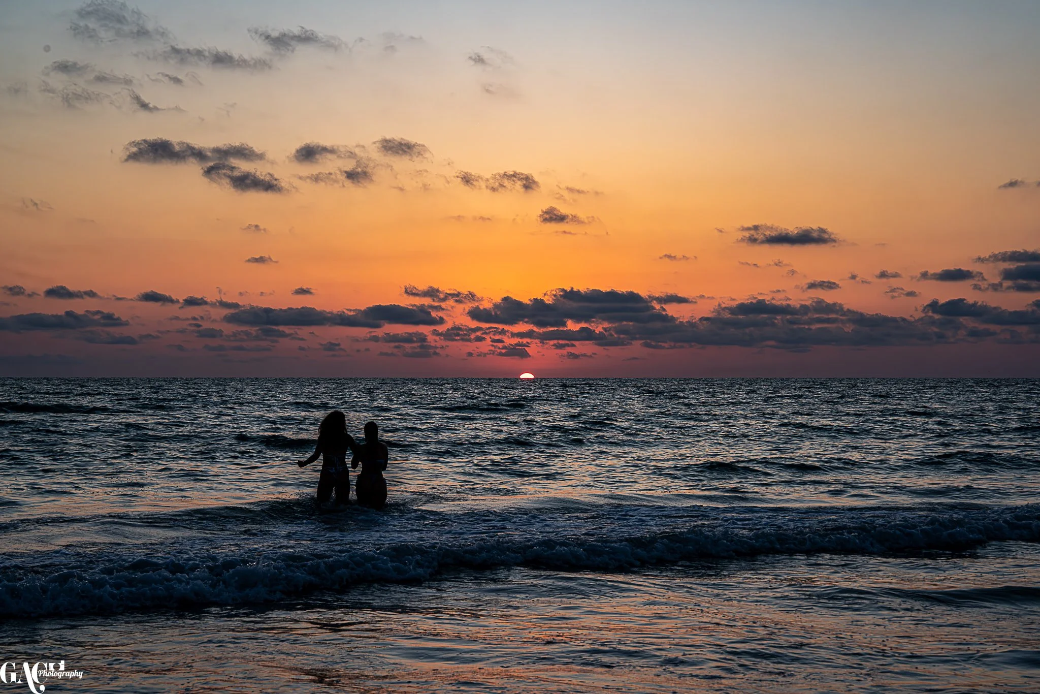 Two women standing in the ocean at sunset, with colorful sky and scattered clouds.