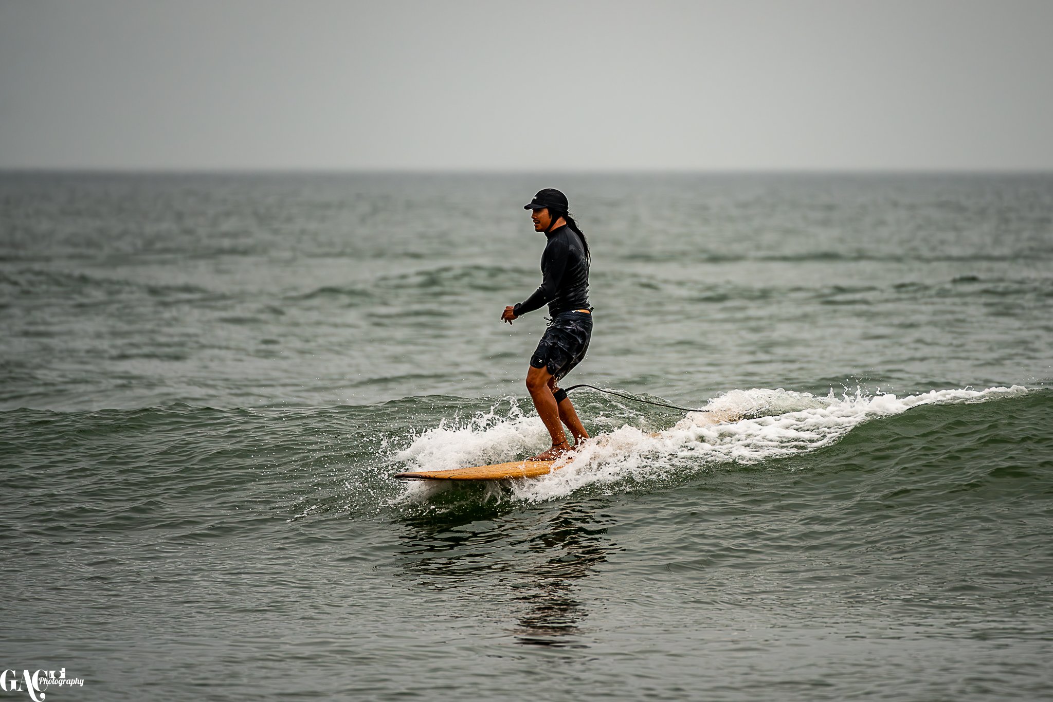 Person wearing black clothing and helmet surfing on a wave with cloudy sky in the background.