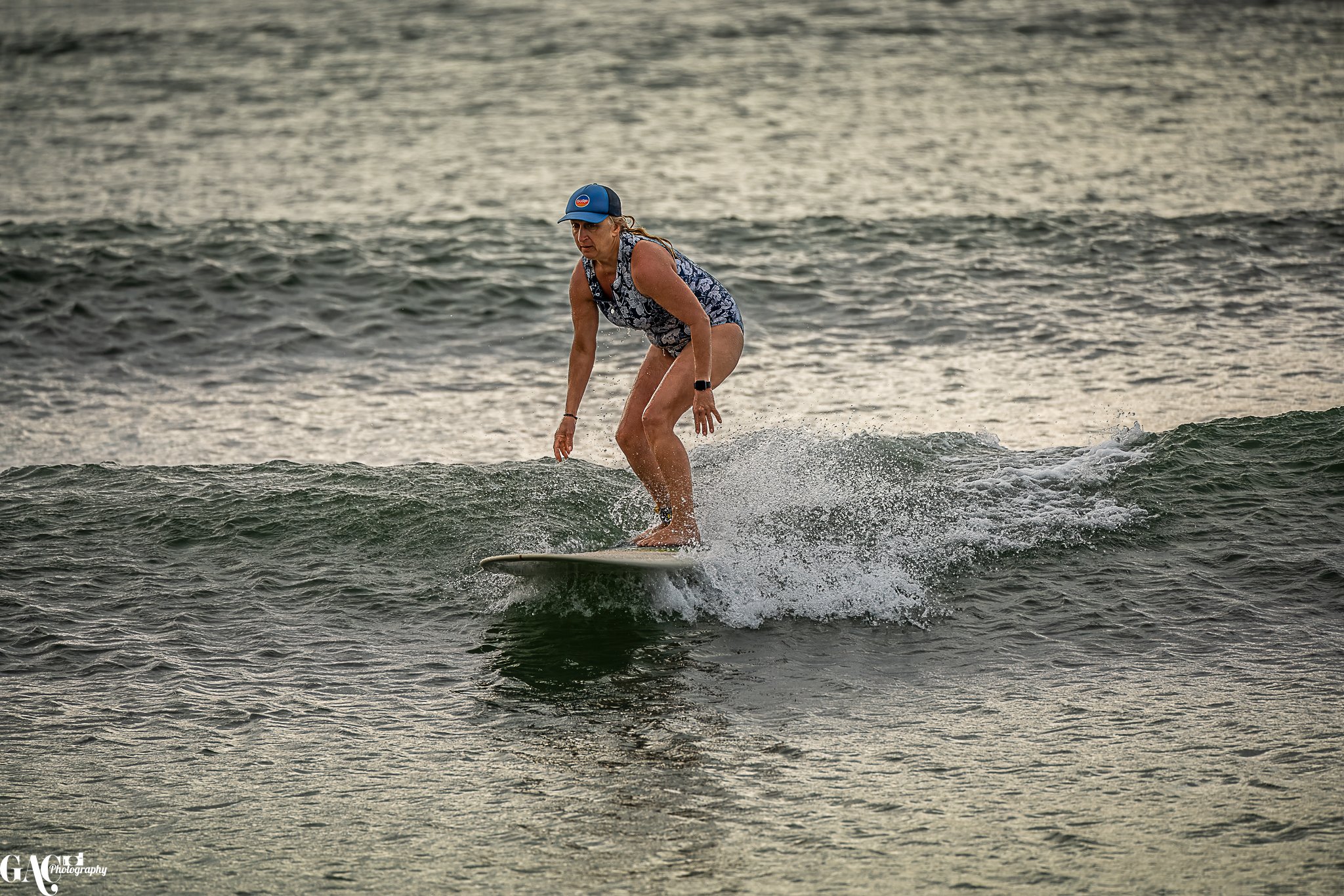 A woman wearing a blue cap and swimsuit rides a small wave on a surfboard in the ocean.