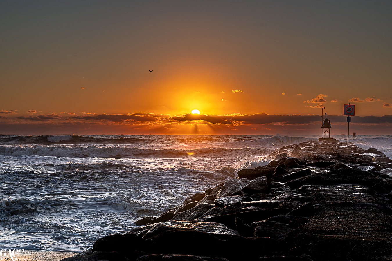 Sunset over the ocean with waves crashing against a rocky jetty, a sign and a small lighthouse structure at the end of the jetty, and a bird flying in the sky.