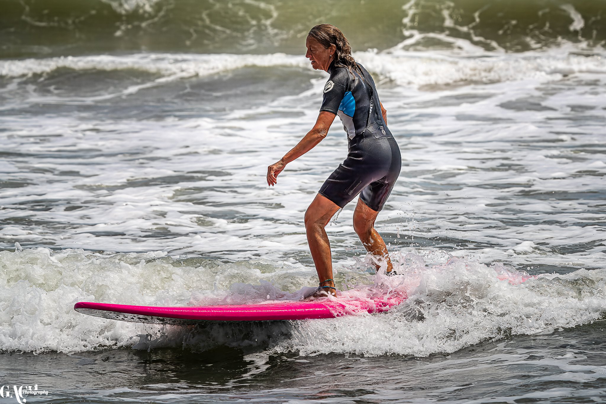 An older woman in a wetsuit surfing on a pink surfboard in the ocean waves.