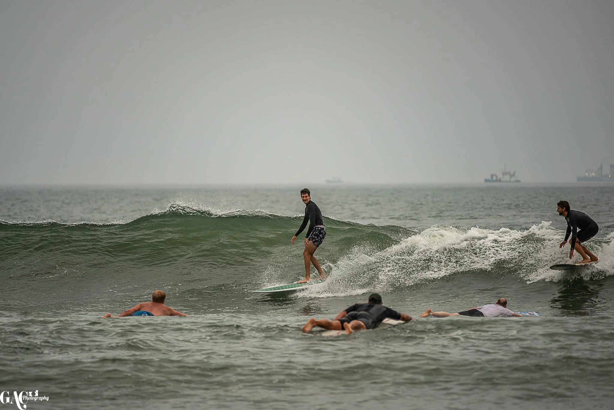 A group of surfers in the ocean, some riding the waves and others lying on their surfboards or swimming.