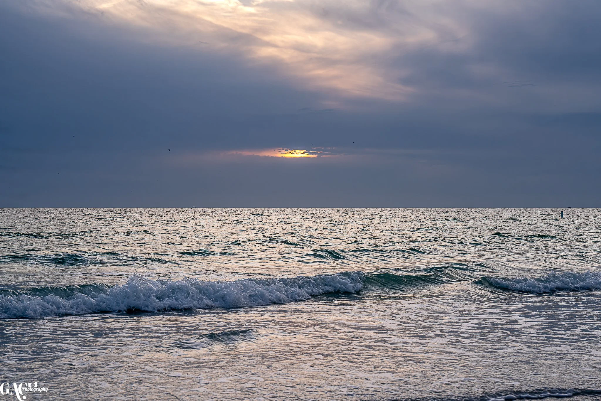 Sunset over a calm ocean with gentle waves and a cloudy sky.