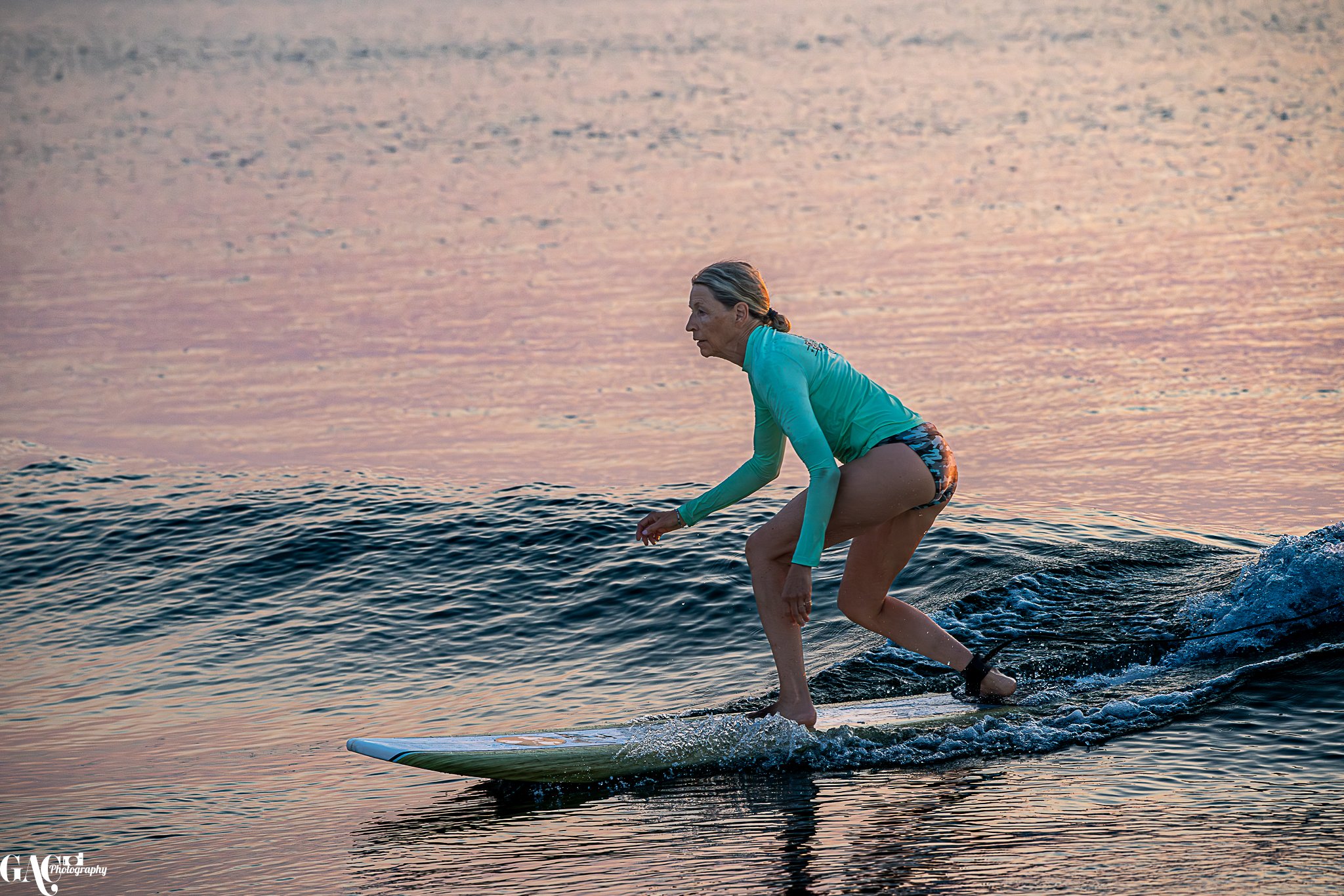 An older woman with blonde hair in a ponytail surfing on a small wave in the ocean during sunset, wearing a turquoise long sleeve shirt and colorful shorts.