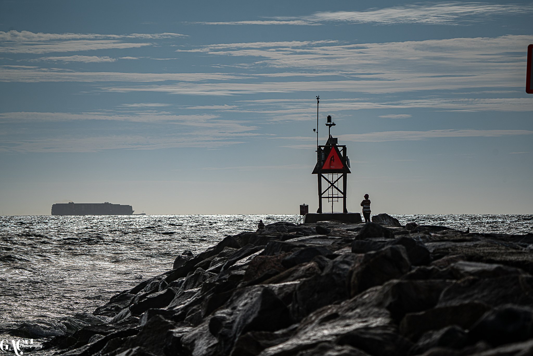 A person standing on rocks at a shoreline next to a small navigational structure with a red warning sign, with the ocean and a large ship in the background under a partly cloudy sky.