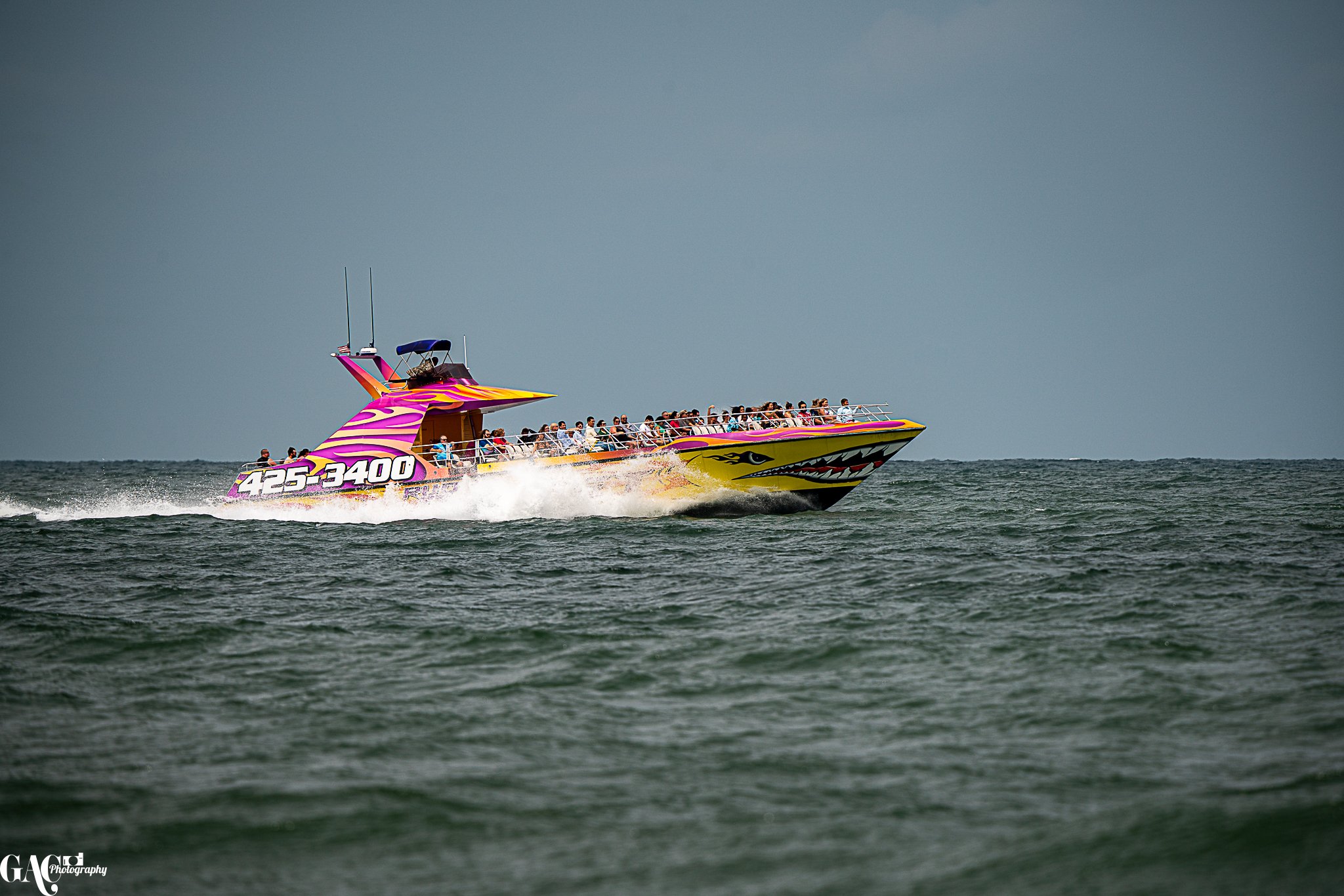 A large speedboat with a colorful dragon face design on the front, colorful purple and yellow pattern on the top, and a phone number "425-3400" on the side. The boat is carrying many passengers and is moving swiftly across the water.