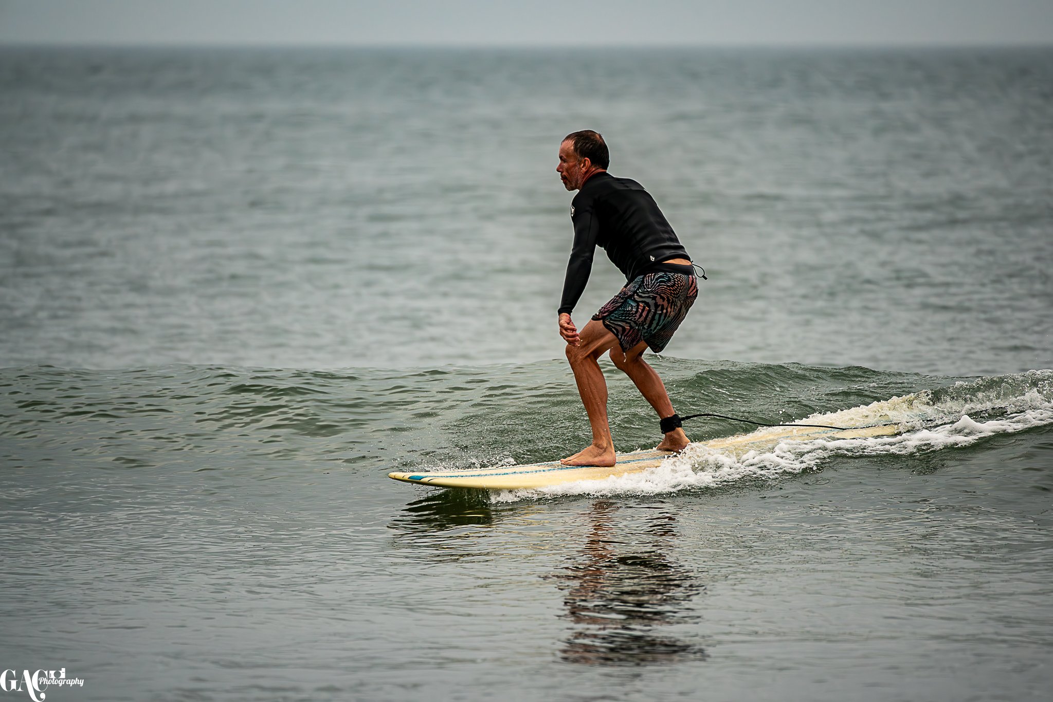 A man surfing on a wave in the ocean, wearing black rash guard and colorful swim shorts.