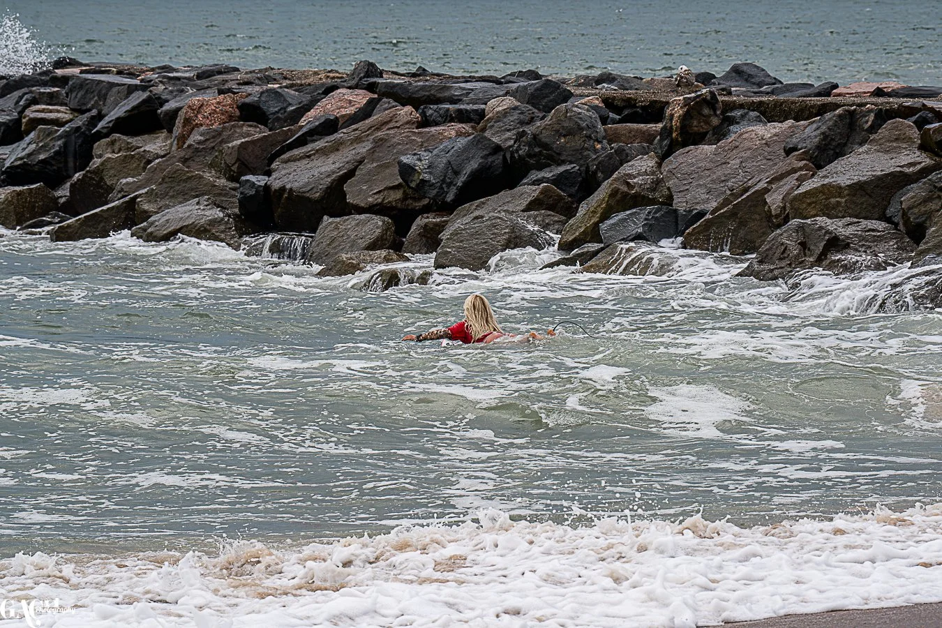 A person in a red wetsuit swimming in the ocean near a rocky jetty.
