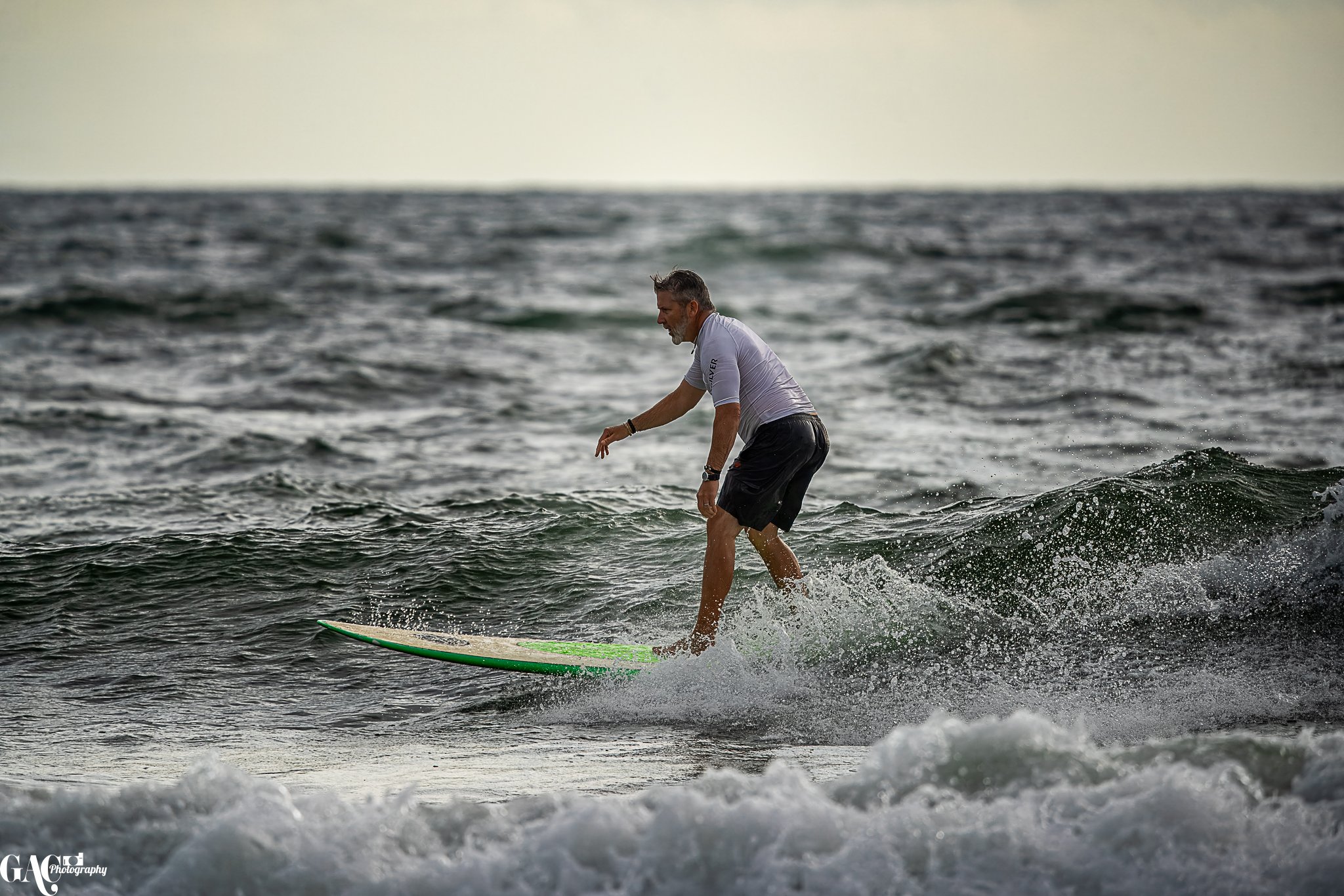 A man with gray hair wearing a white t-shirt and black shorts surfing on a green surfboard in the ocean.