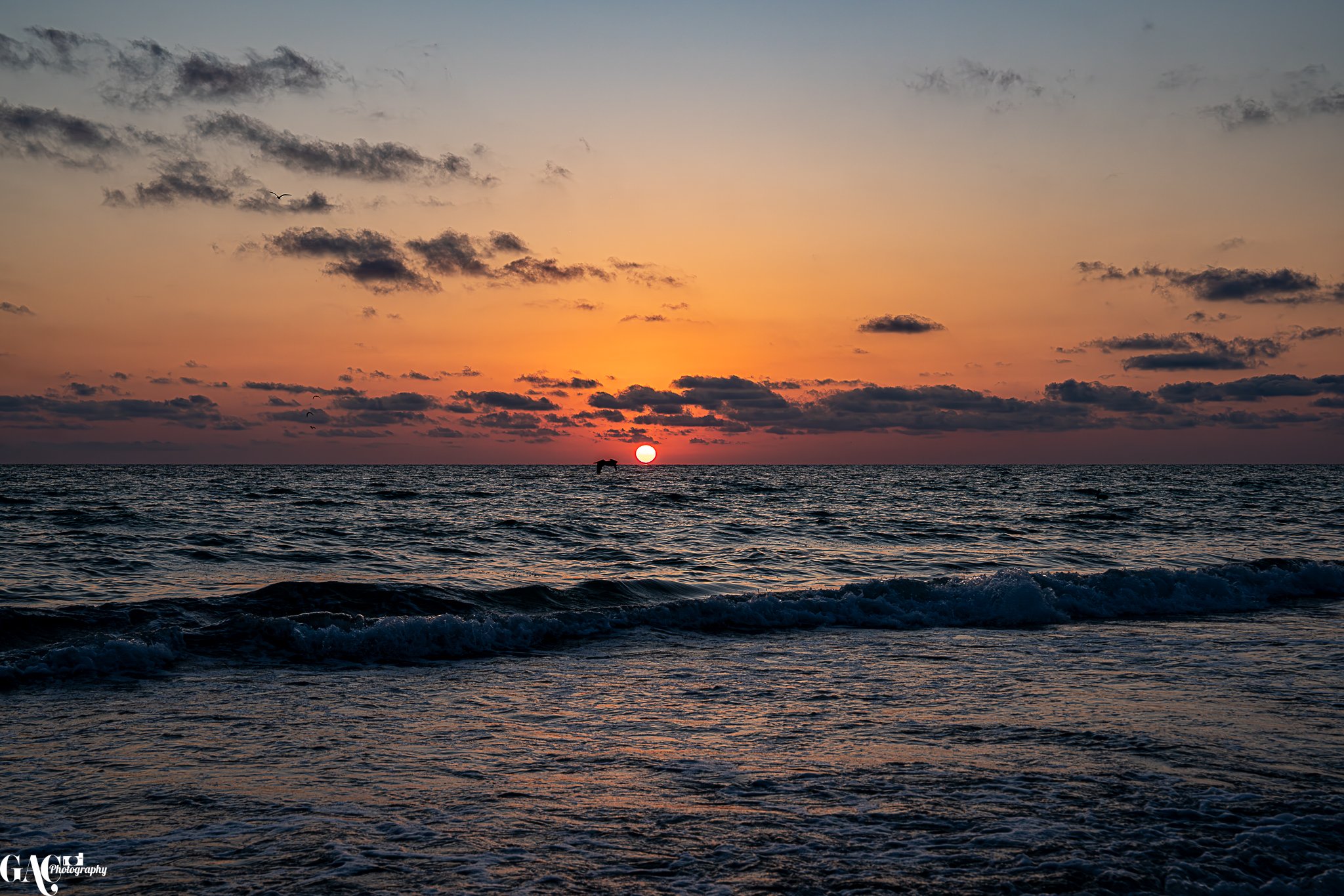 Ocean sunset with sun on horizon, scattered clouds, and gentle waves.