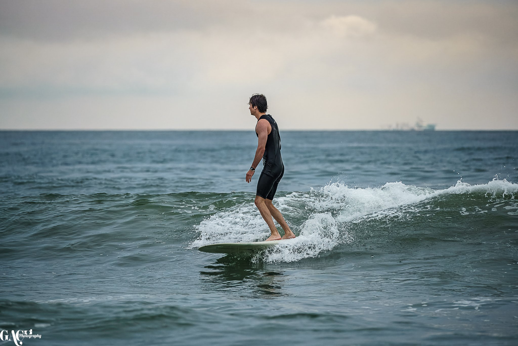 A man wearing a black wetsuit surfing on a small wave in the ocean, with a cargo ship visible in the distance under a cloudy sky.