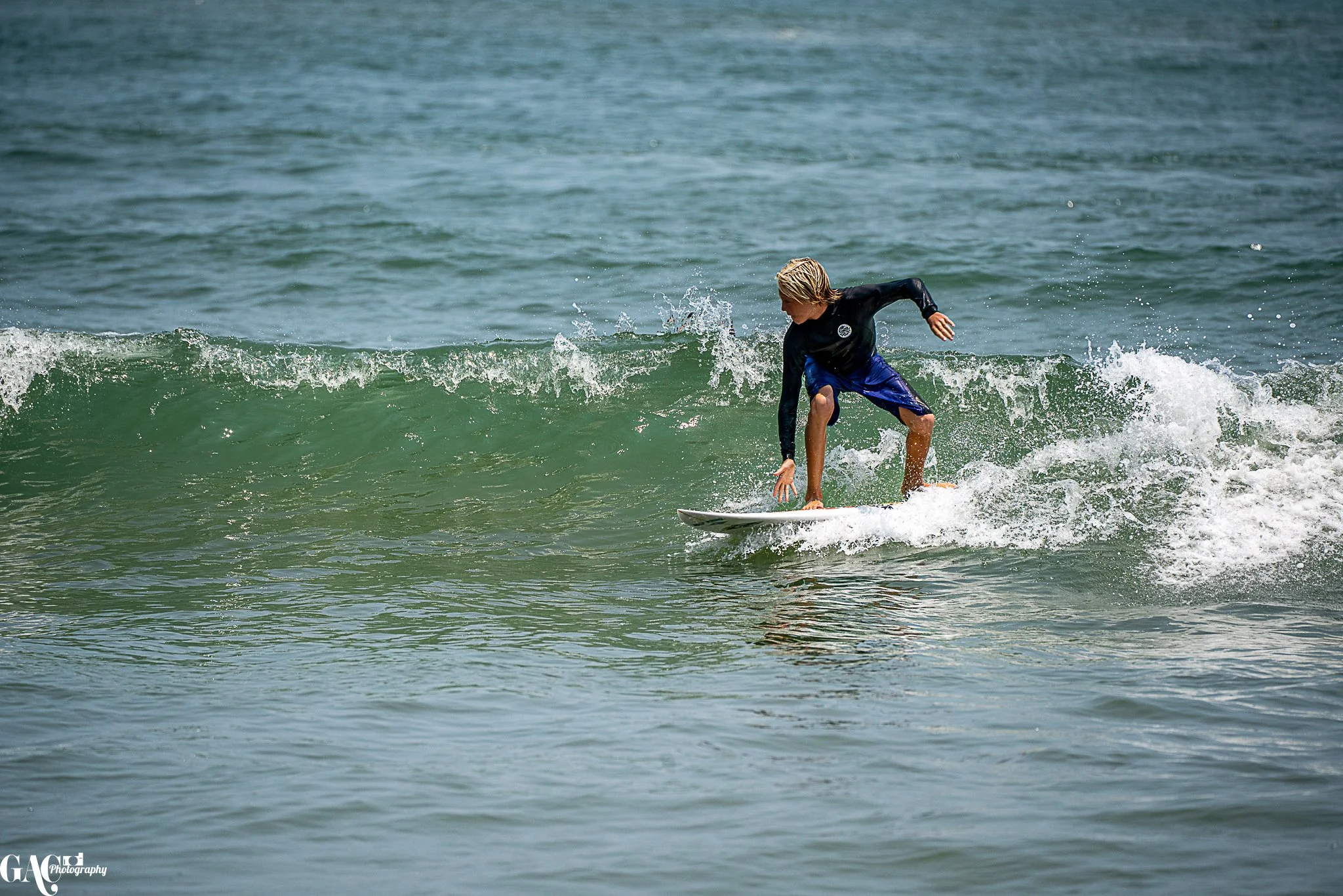 A young boy with blond hair surfing on a small wave in the ocean, wearing a black long-sleeve rash guard and blue swim shorts.