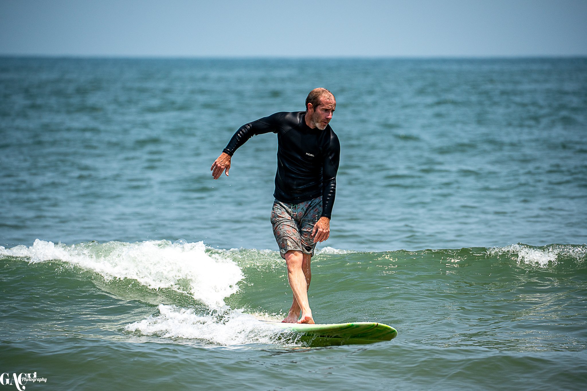 A man with gray hair and beard surfing on a green surfboard in the ocean, wearing a black long-sleeve rash guard and patterned shorts, with a blue sky in the background.