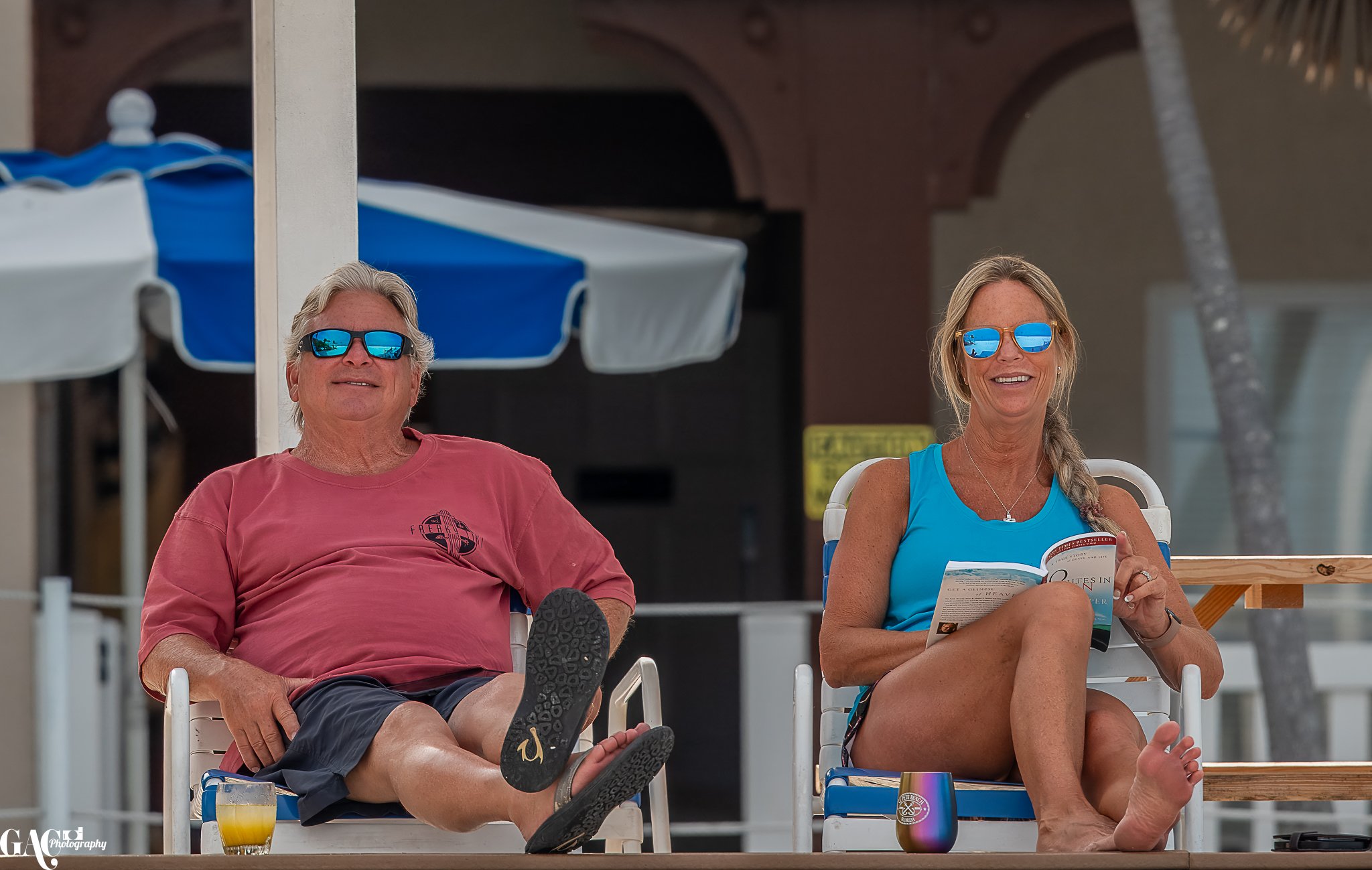Two people relaxing on lounge chairs wearing sunglasses, outdoors with beach umbrellas in the background.