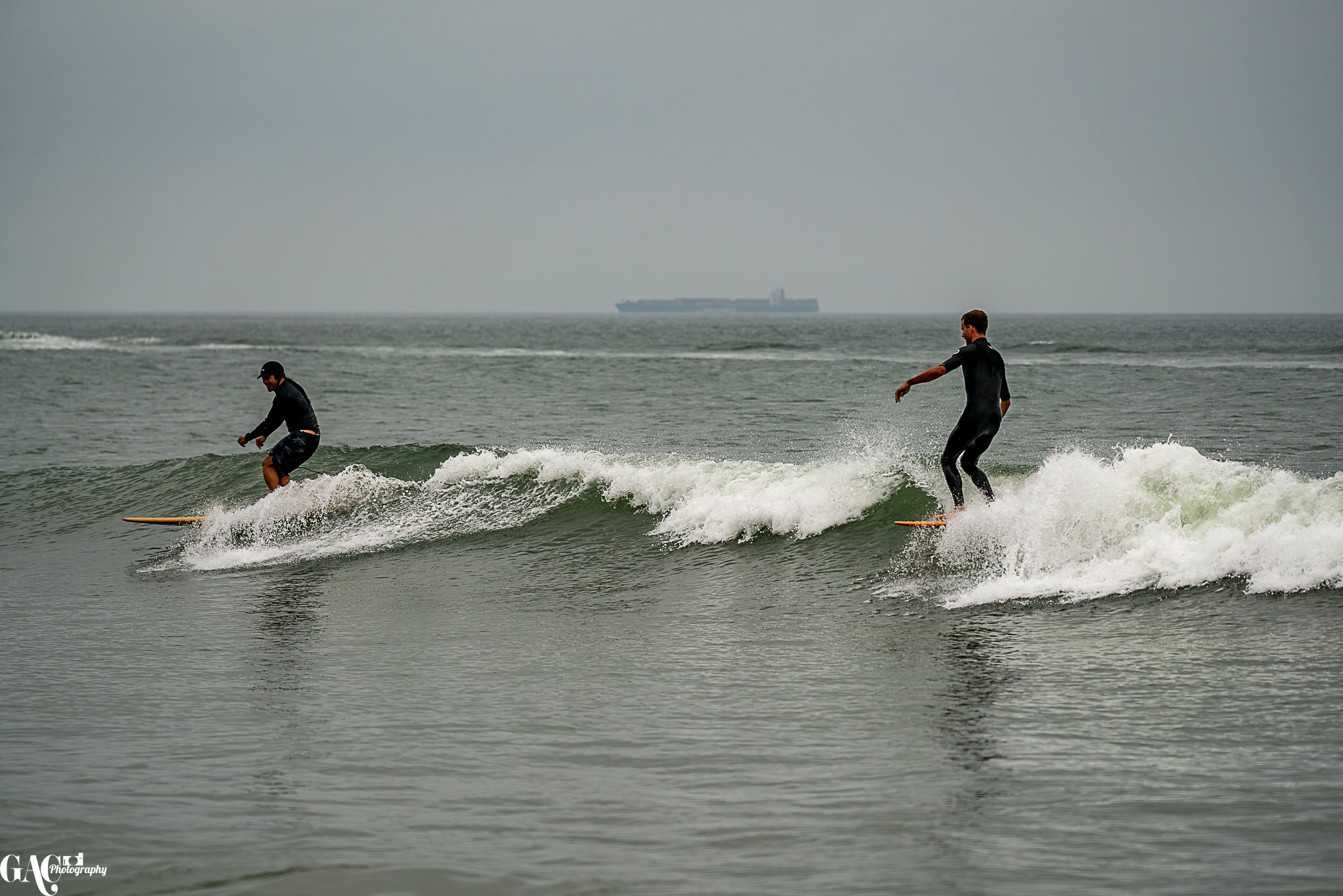 Two surfers riding small waves at the beach with a cargo ship in the distant background on a cloudy day.