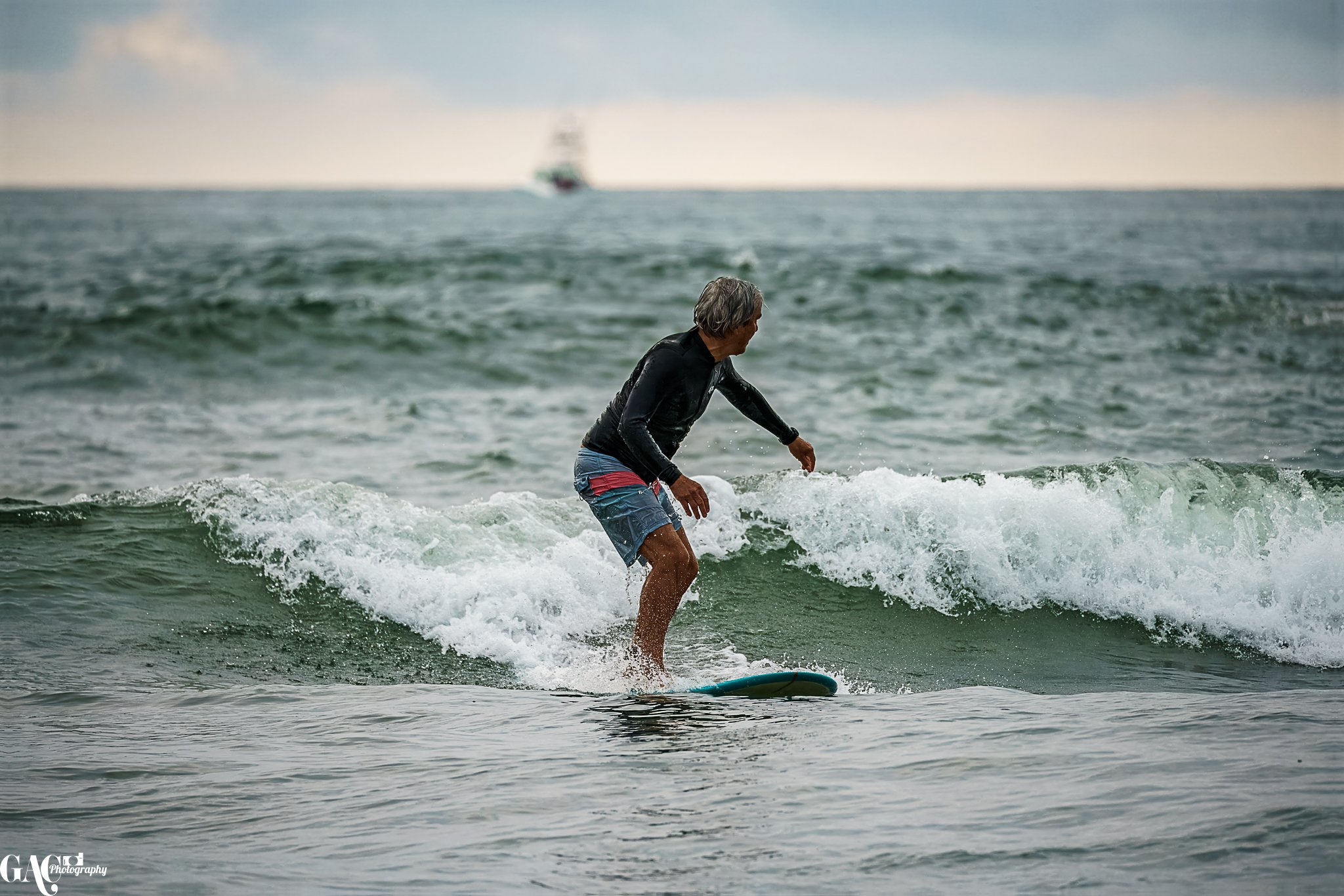 An older man surfing on a small wave in the ocean, wearing a black wetsuit and colorful swim trunks, with a boat in the distance.