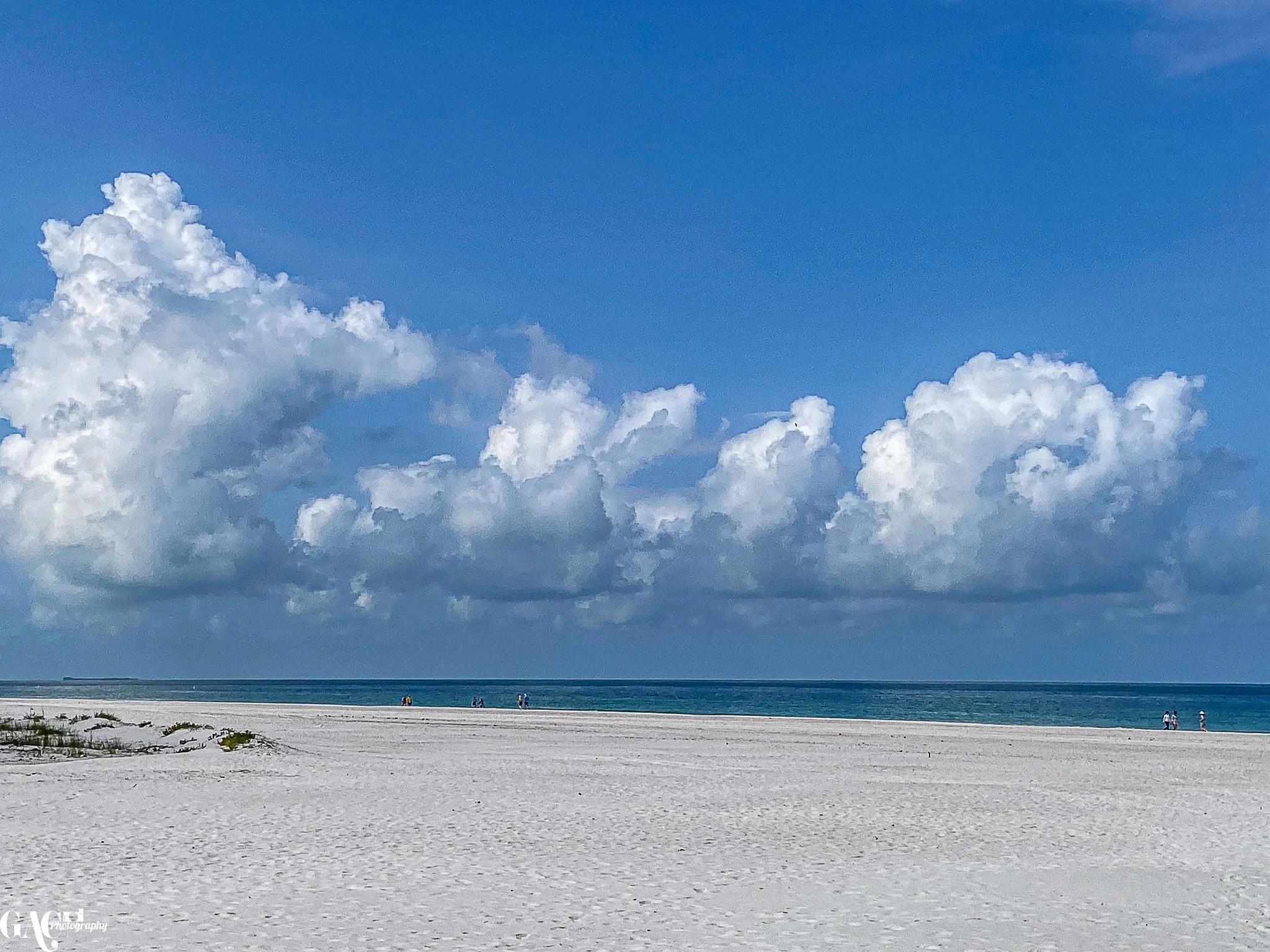 Empty sandy beach with a few people walking along the shoreline under a blue sky with large white clouds.