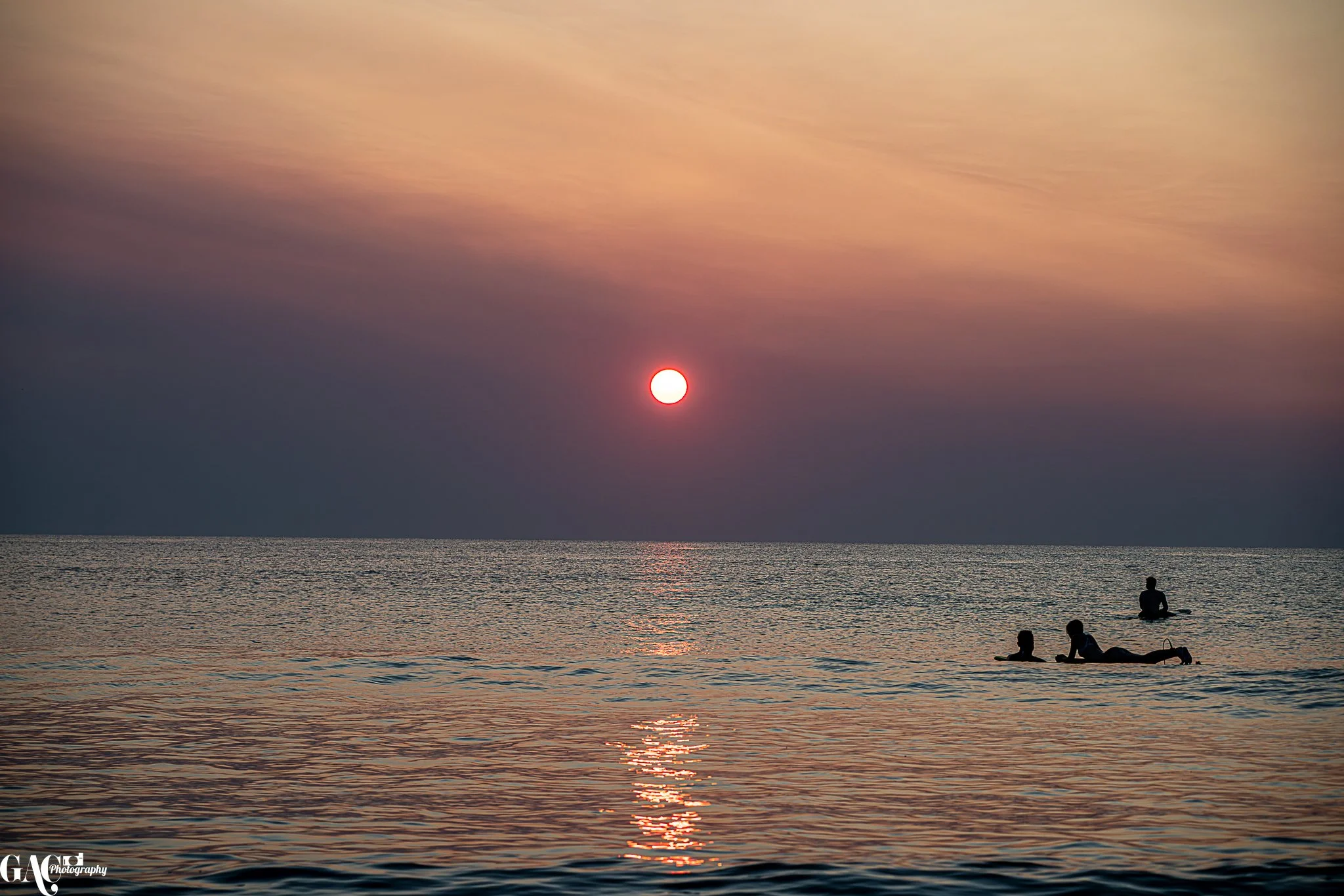 Silhouettes of children on paddleboards in the ocean during sunset, with a colorful sky and the sun near the horizon.