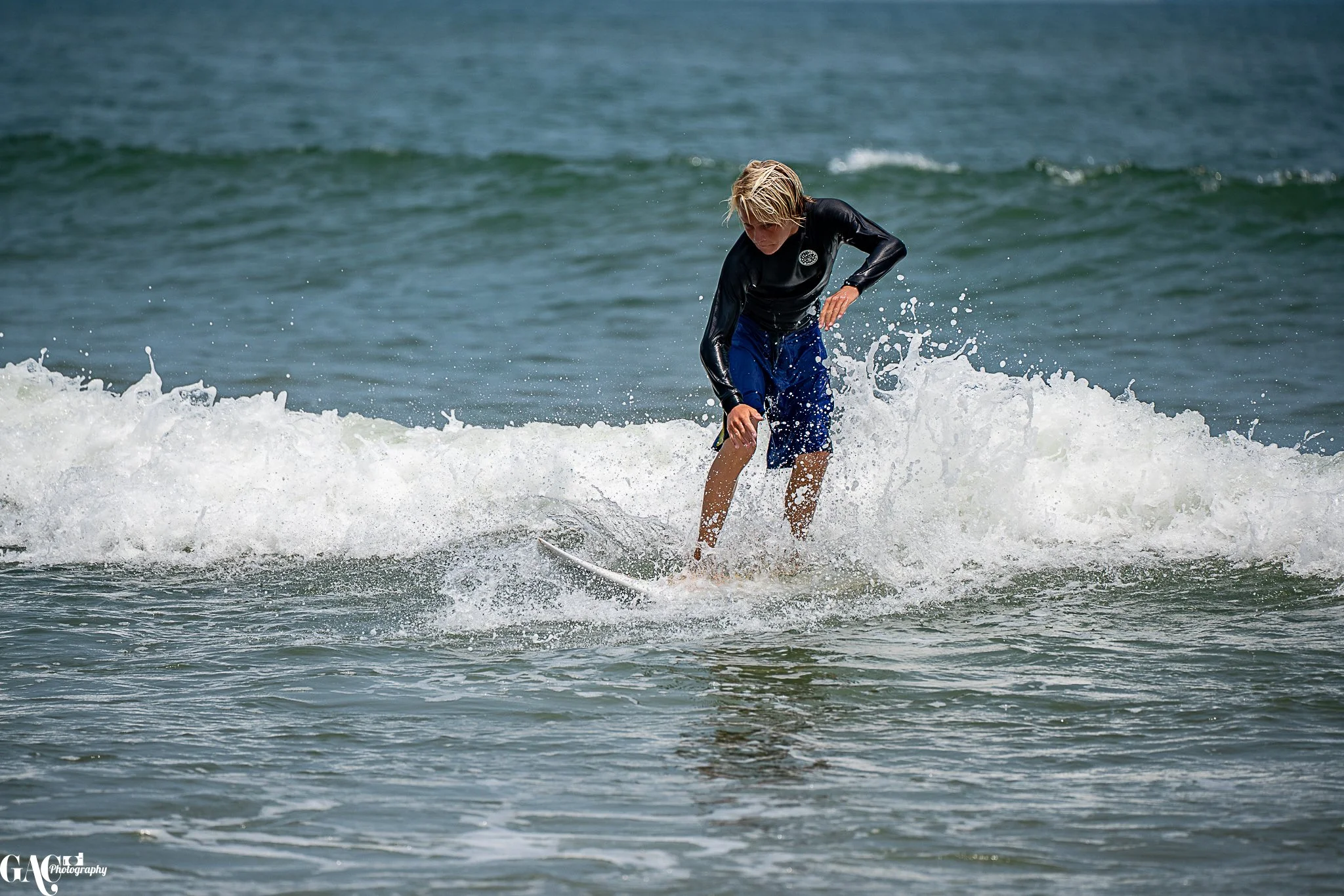 A person surfing on a small wave in the ocean, wearing a black wetsuit and blue shorts.