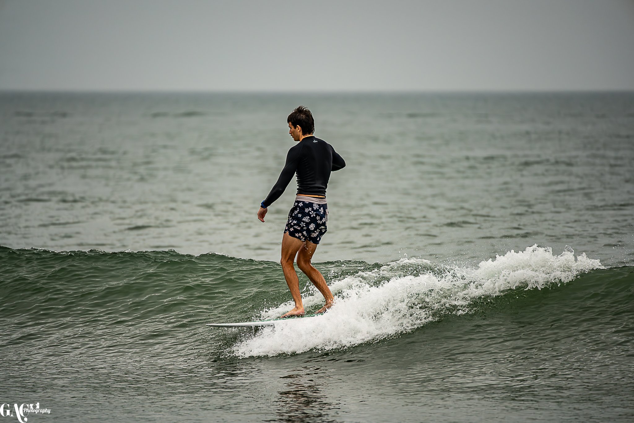 A man surfing on a small wave in the ocean, wearing black rash guard and floral swim trunks, on a cloudy day.
