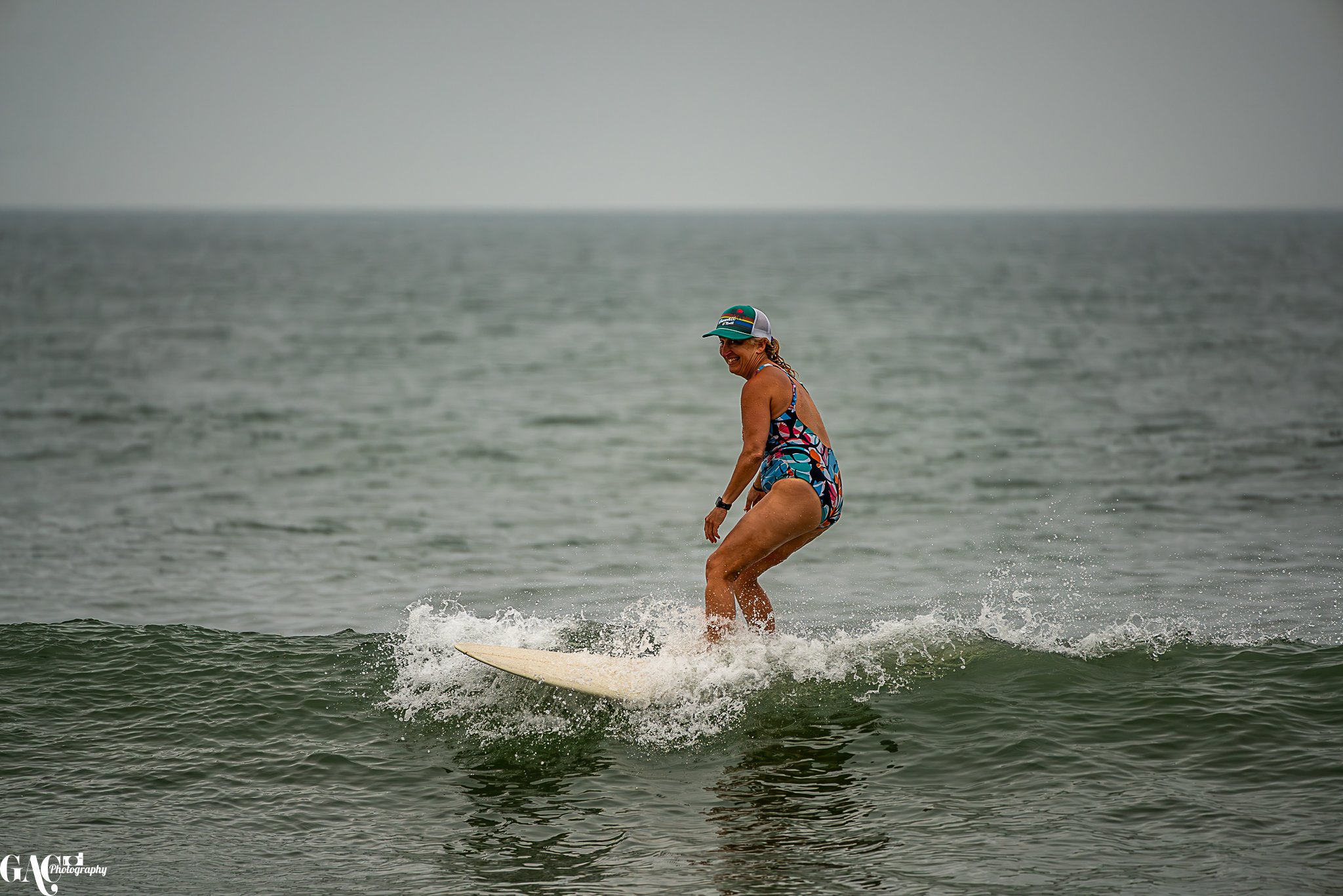 A woman wearing a floral swimsuit and a cap is surfing on a small wave in the ocean, smiling as she rides the wave.