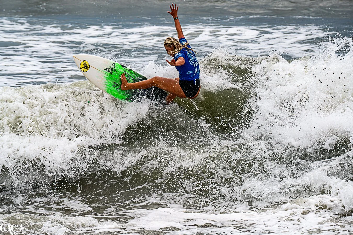 A woman surfing on a wave with a green and white surfboard, wearing a blue shirt and black shorts at the ocean.