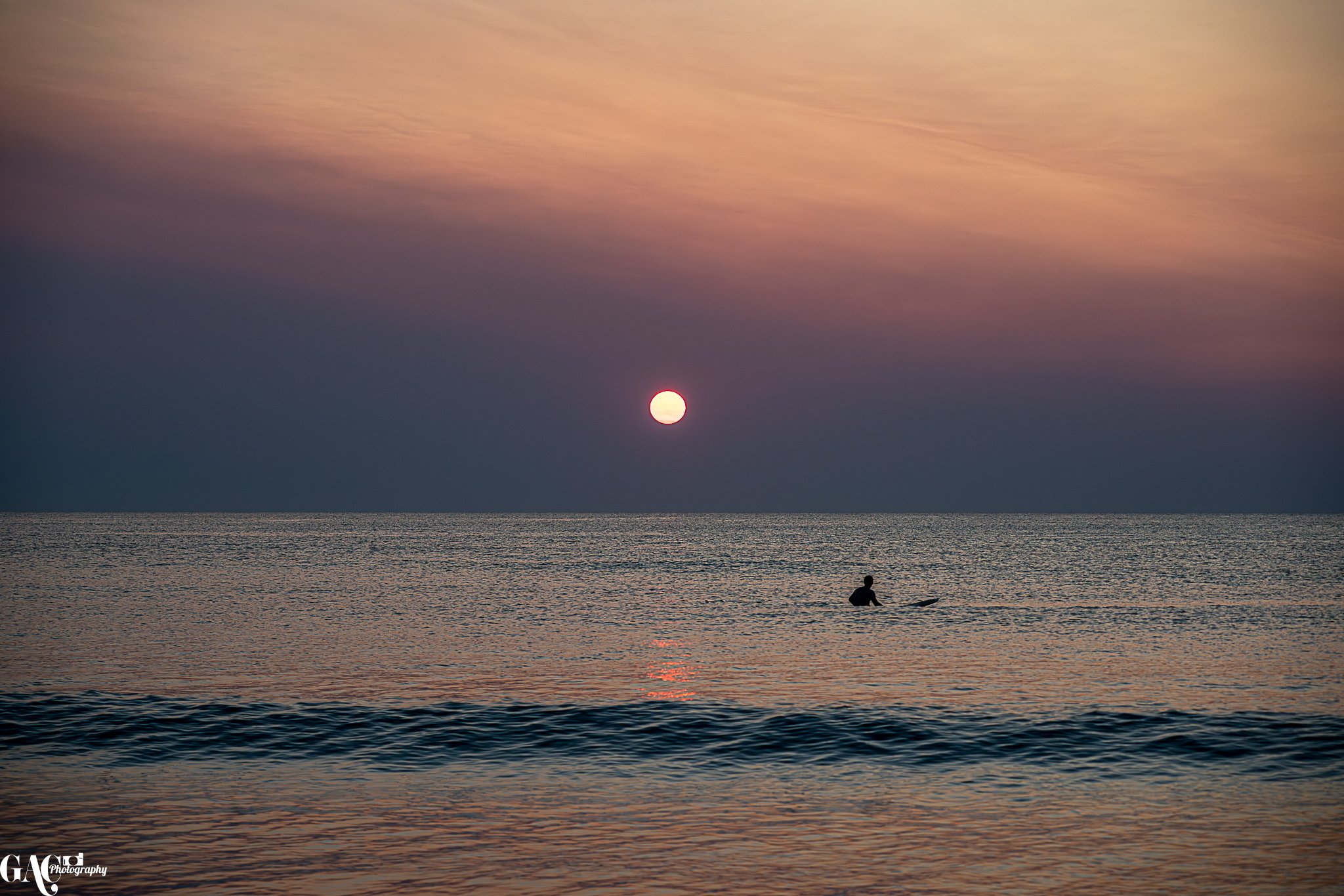 Silhouette of a person surfing in the ocean during sunset, with a large sun low in the sky and colorful clouds above.
