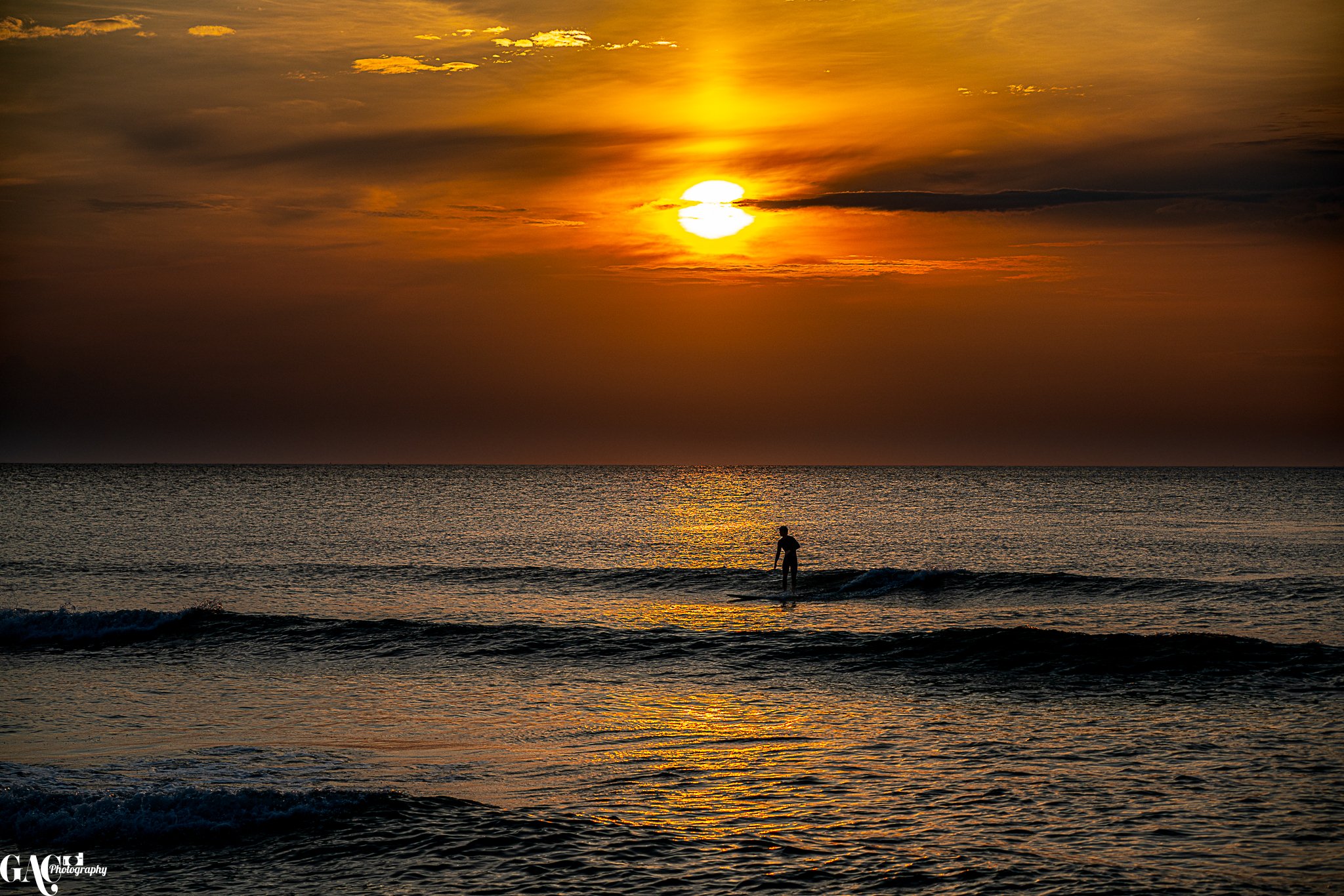 A person standing on a surfboard in the ocean during sunset, with the sun partially covered by clouds and the sky painted in warm orange and yellow hues.