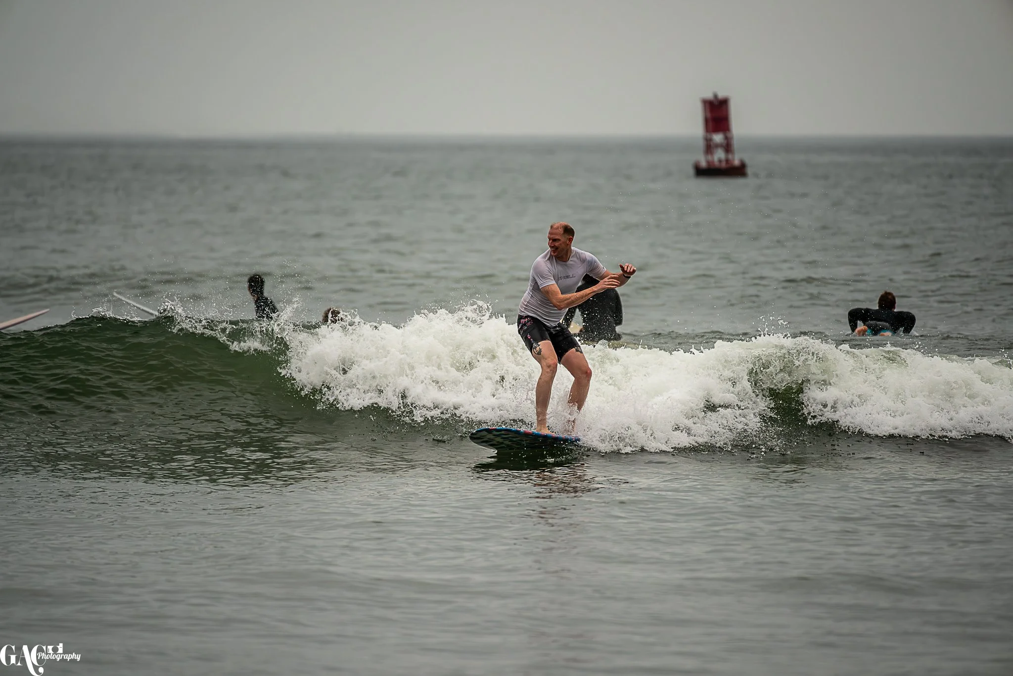 A man riding a surfboard on a wave in the ocean with two other surfers in the background and a red lighthouse in the distance.