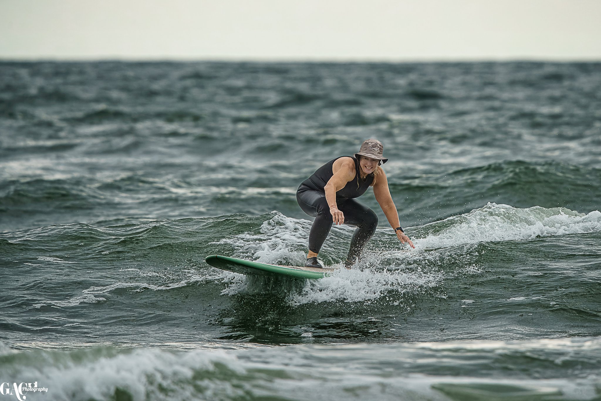 A woman riding a surfboard on the ocean waves, smiling and wearing a hat and black wetsuit.