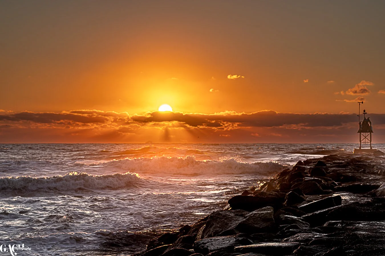 A sunset over the ocean with waves crashing against rocks and a small lighthouse on the rocky shore, glowing in the warm light.