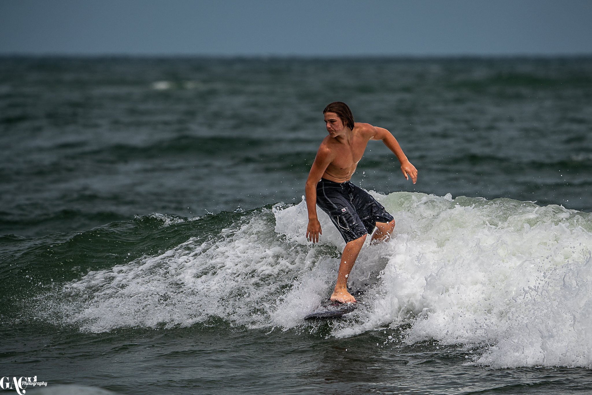 A shirtless man with long hair wearing black shorts riding a surfboard on a small wave in the ocean.