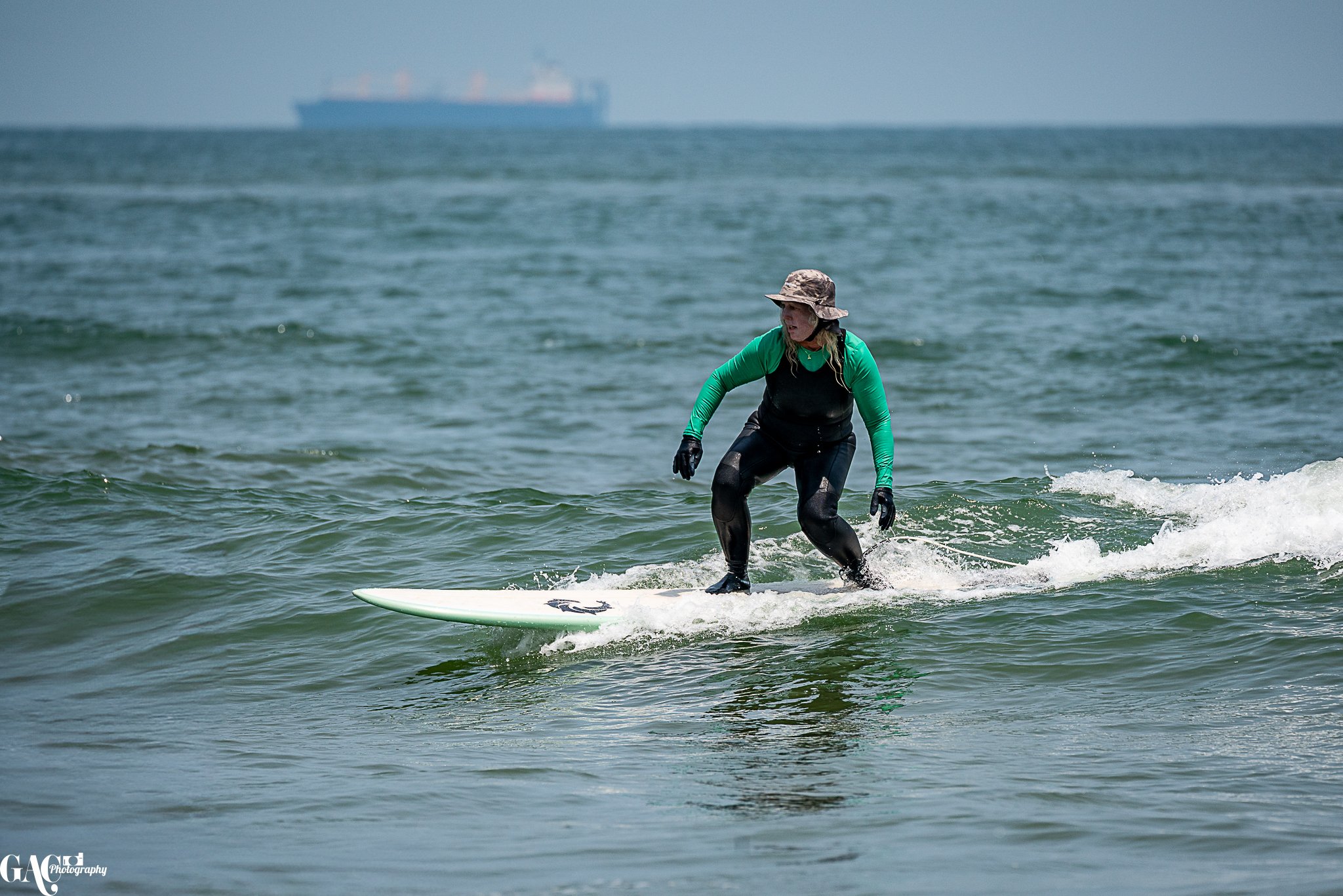 A woman surfing on a wave in the ocean, wearing a camouflage hat, green long-sleeve shirt, black vest, black wetsuit and gloves, with a large cargo ship in the background.