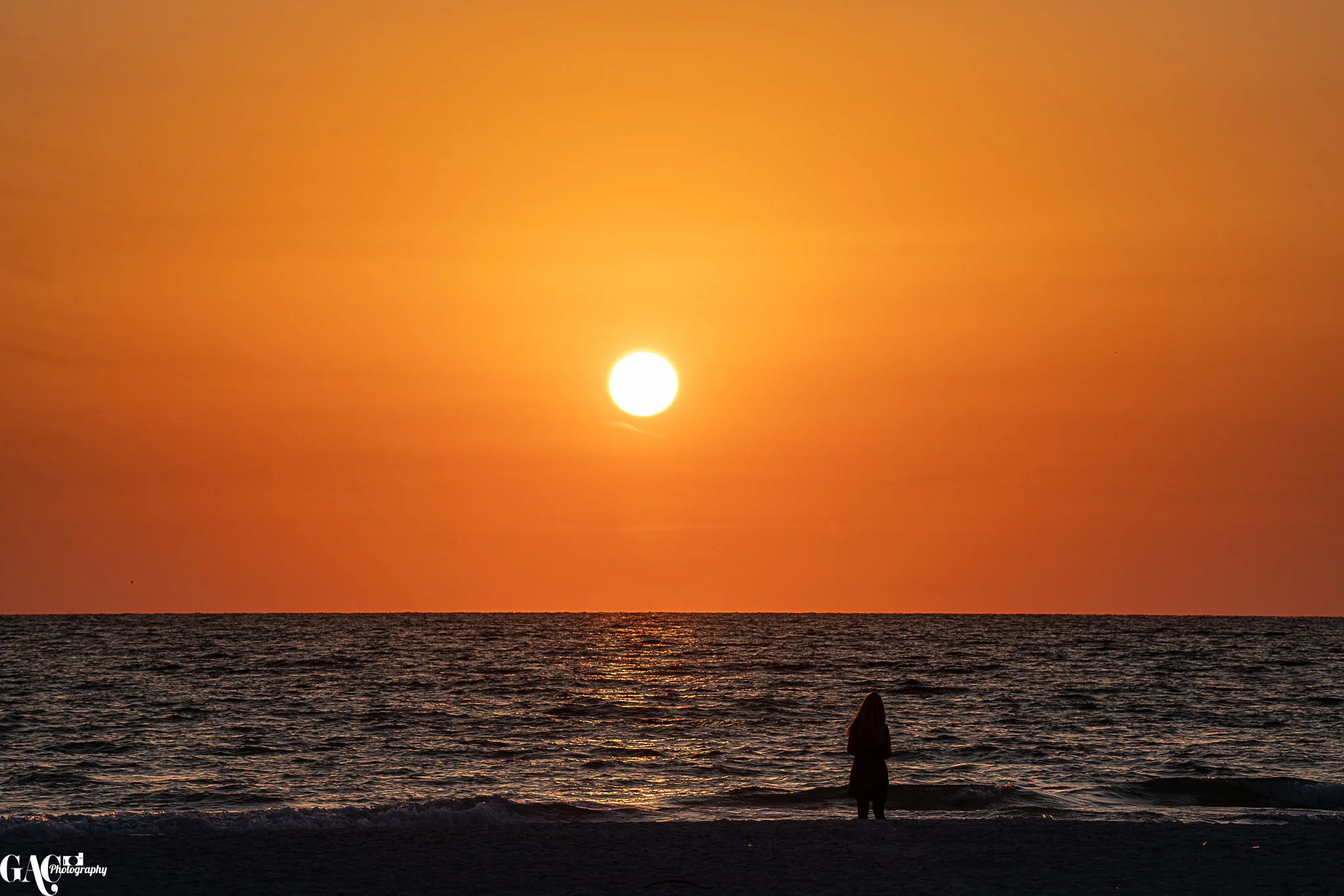 A person standing on the beach at sunset, facing the ocean with the sun low on the horizon and the sky painted in orange and yellow hues.