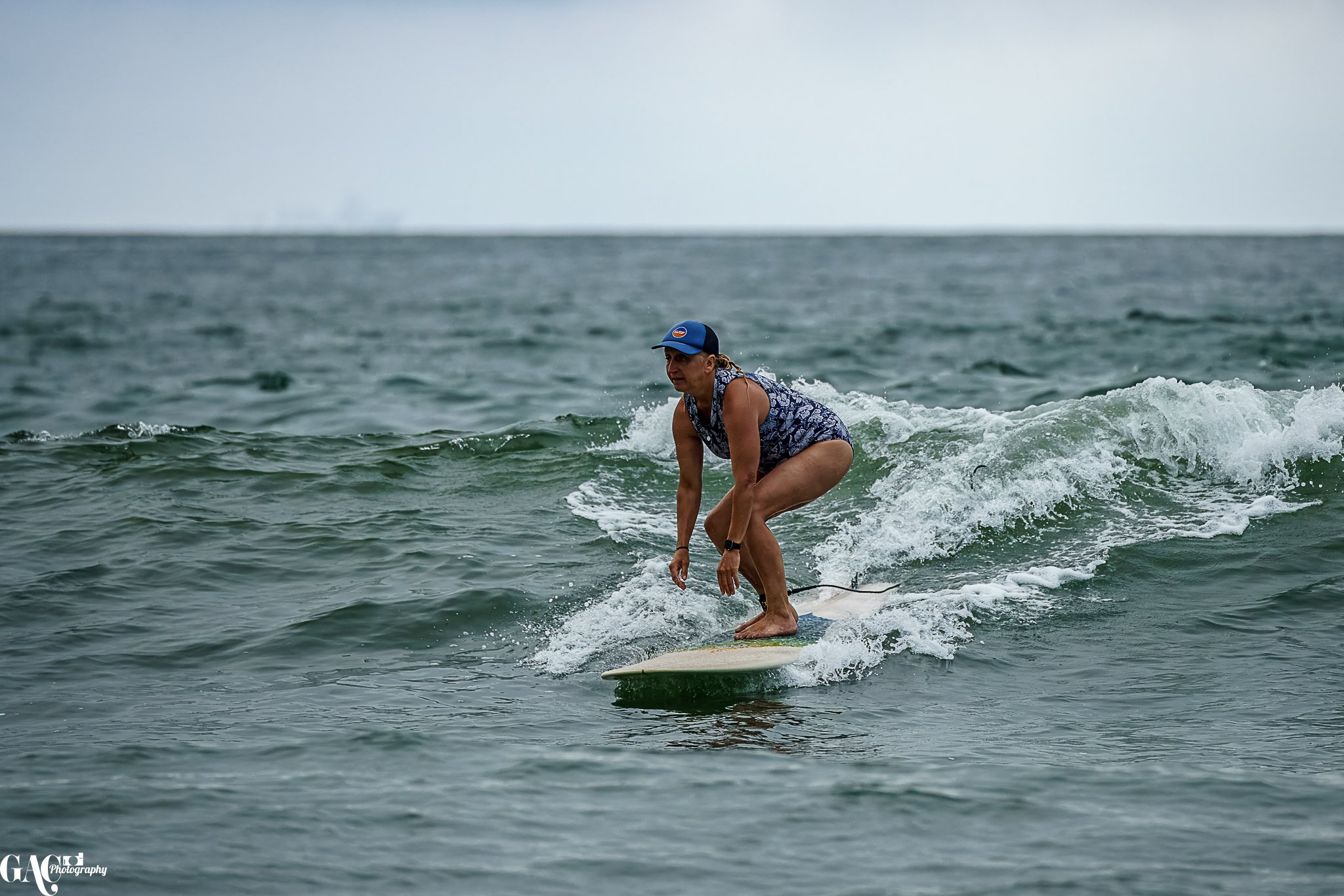 A woman surfing on a small wave in the ocean, wearing a blue cap, a sleeveless top, and shorts.