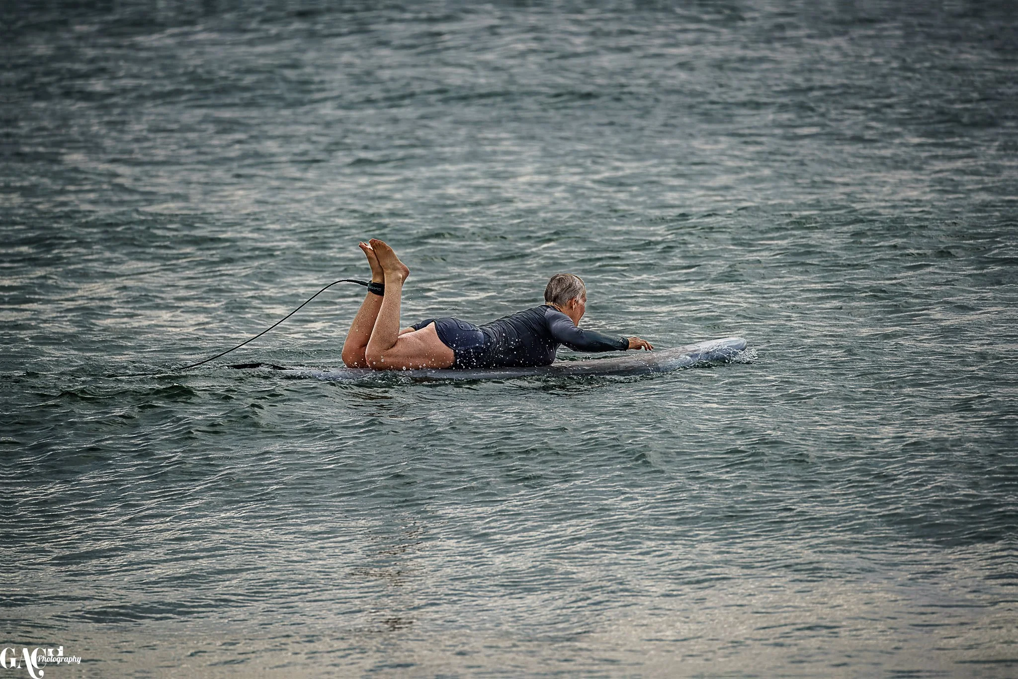 A person in a wetsuit lying on a surfboard in the water, paddling with one arm.