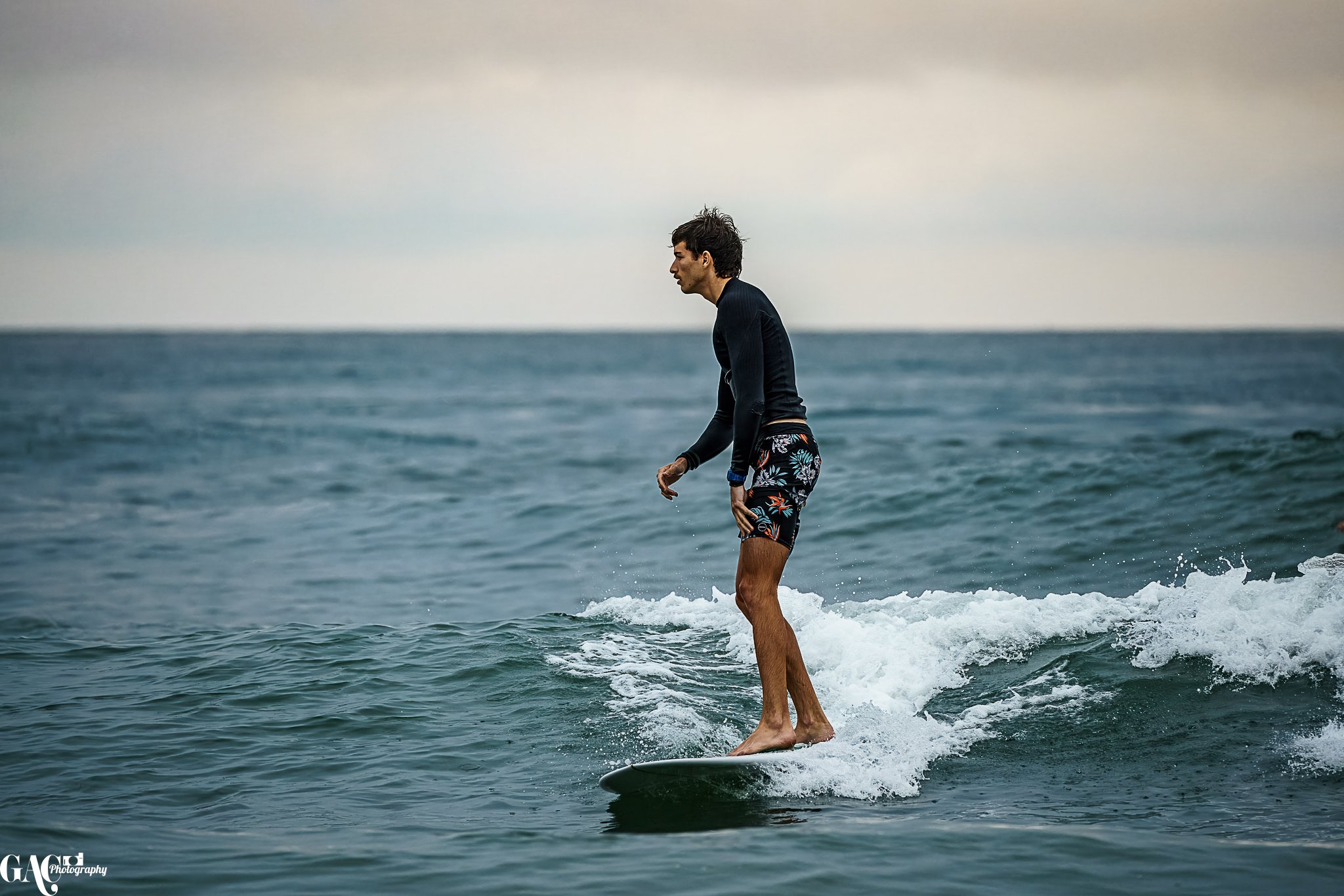 A man surfing on the ocean waves, wearing a black long-sleeve rash guard and colorful floral swim shorts.
