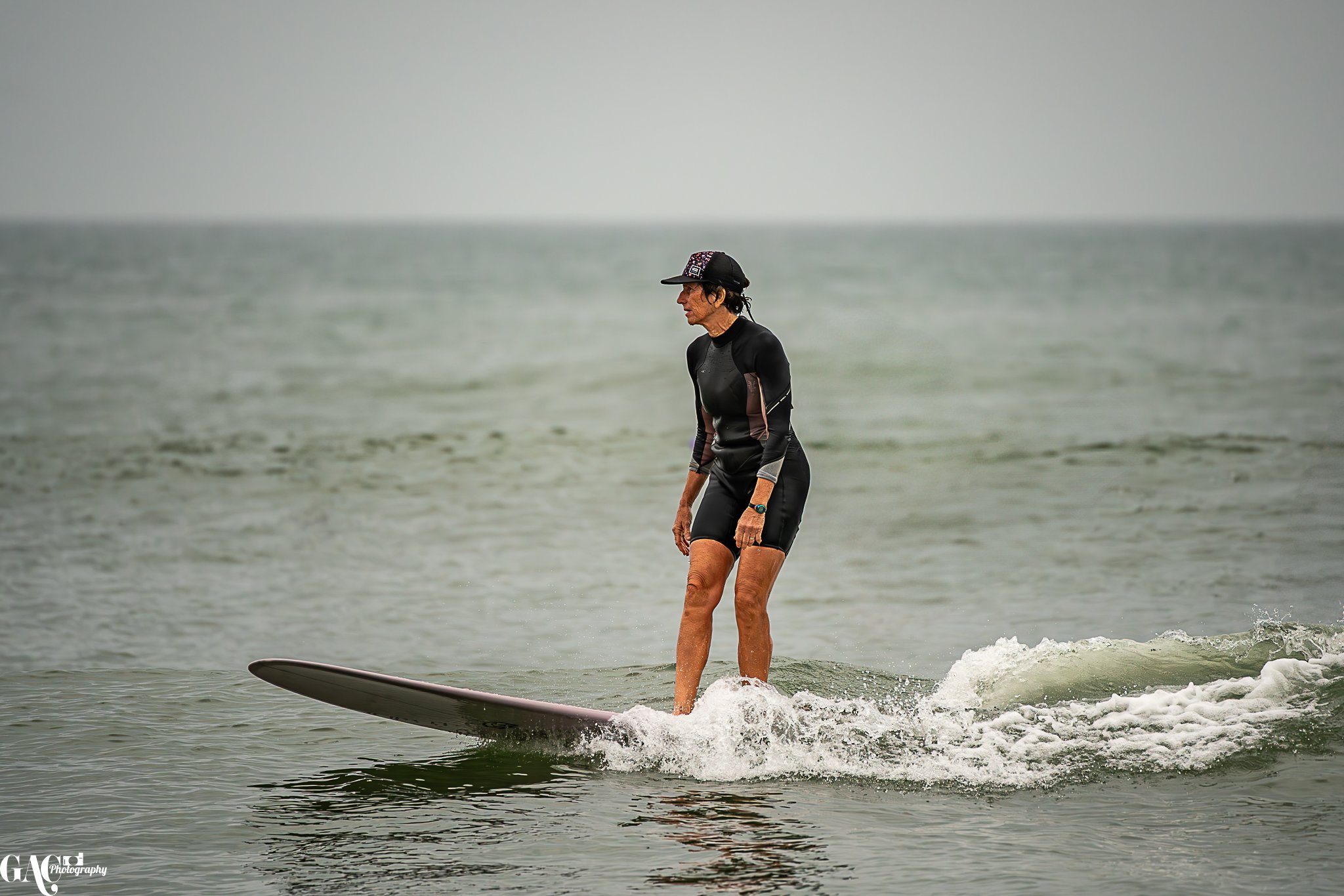 Woman surfing on a wave in the ocean, wearing a black wetsuit and a black cap.