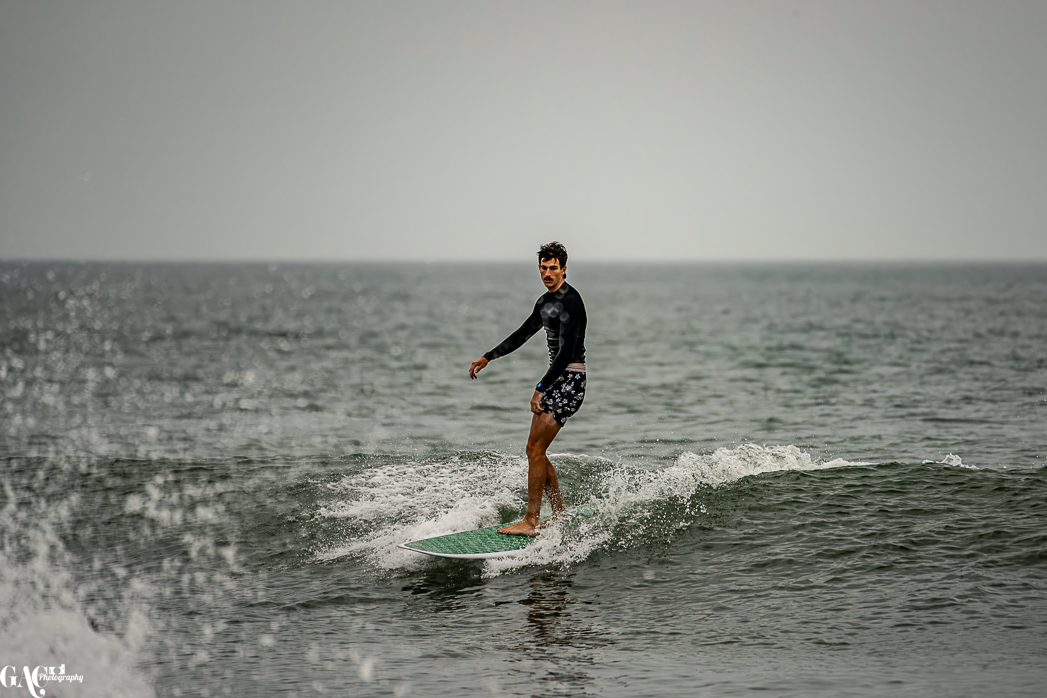 A man surfing on a small wave in the ocean, wearing a black long-sleeve rash guard and floral swim trunks, looking towards the camera.