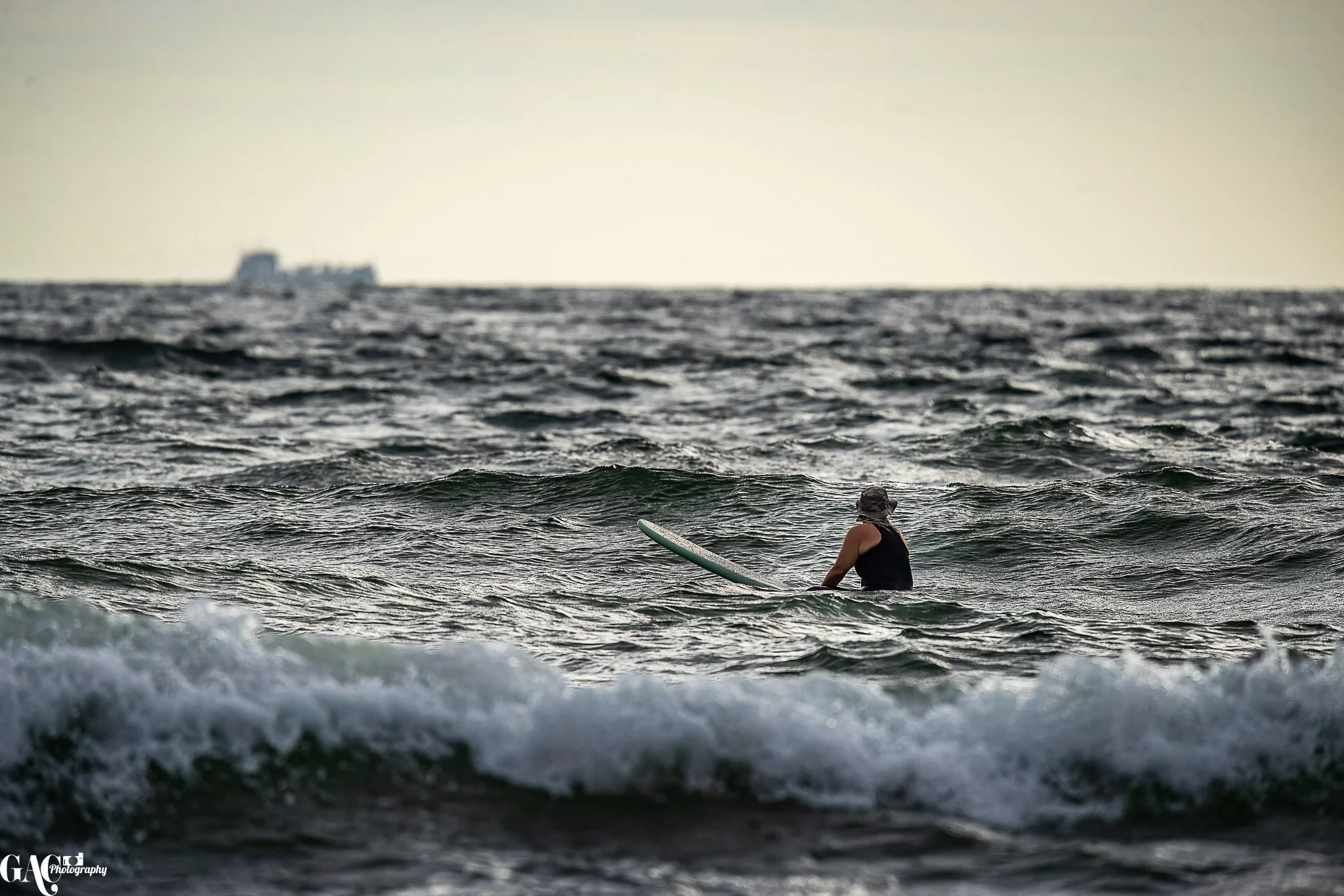 A person in the ocean holding a surfboard, with a ship visible in the distant background.