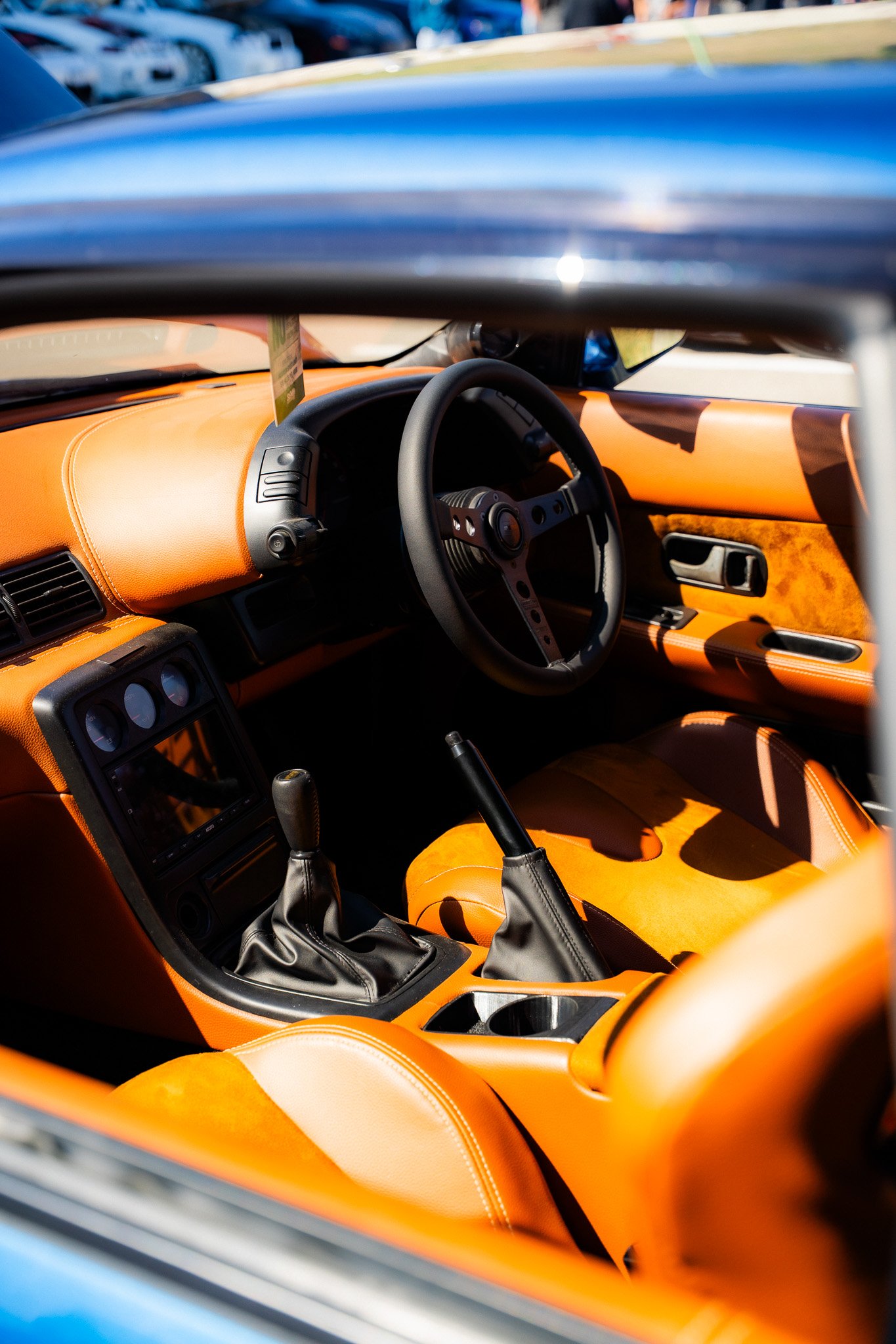 Interior view of a sports car with orange leather interior, black steering wheel, and manual shifter, parked among other cars.