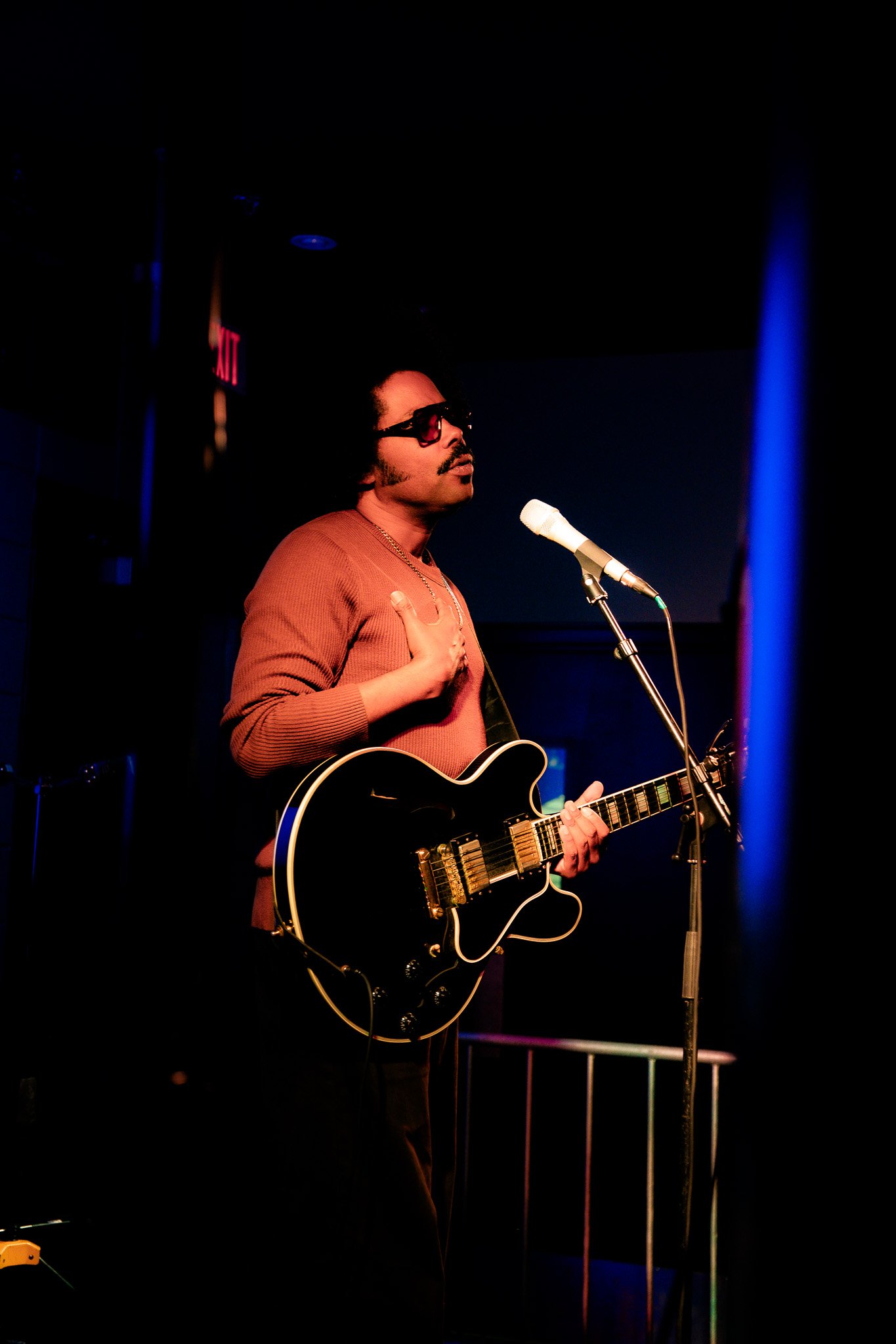 A man wearing sunglasses and a brown long-sleeve shirt playing an electric guitar and singing into a microphone on stage with a dark background.