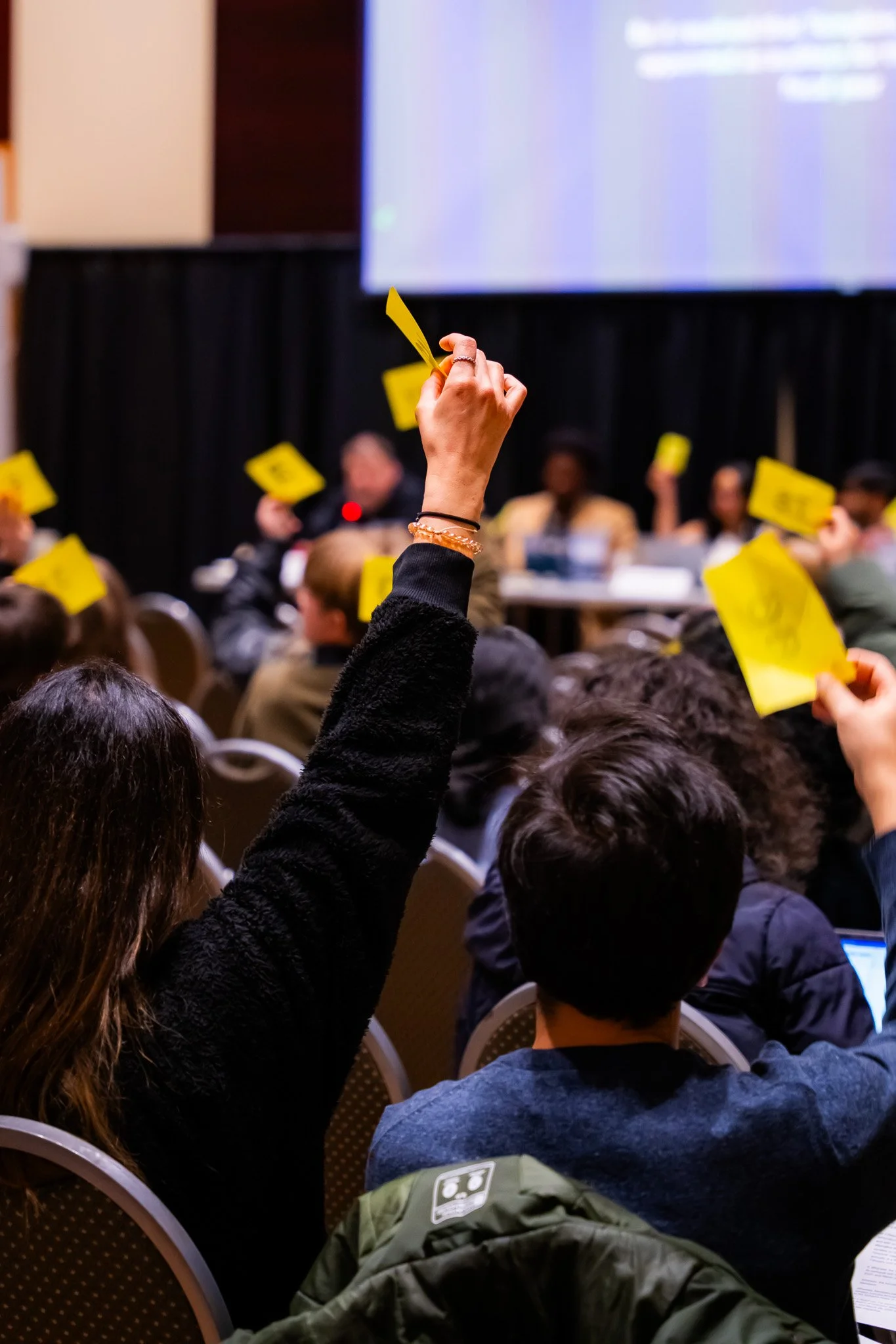 Audience seated at a conference or seminar; a woman raises her hand holding a yellow sticky note; people have notebooks and bags with conference materials.