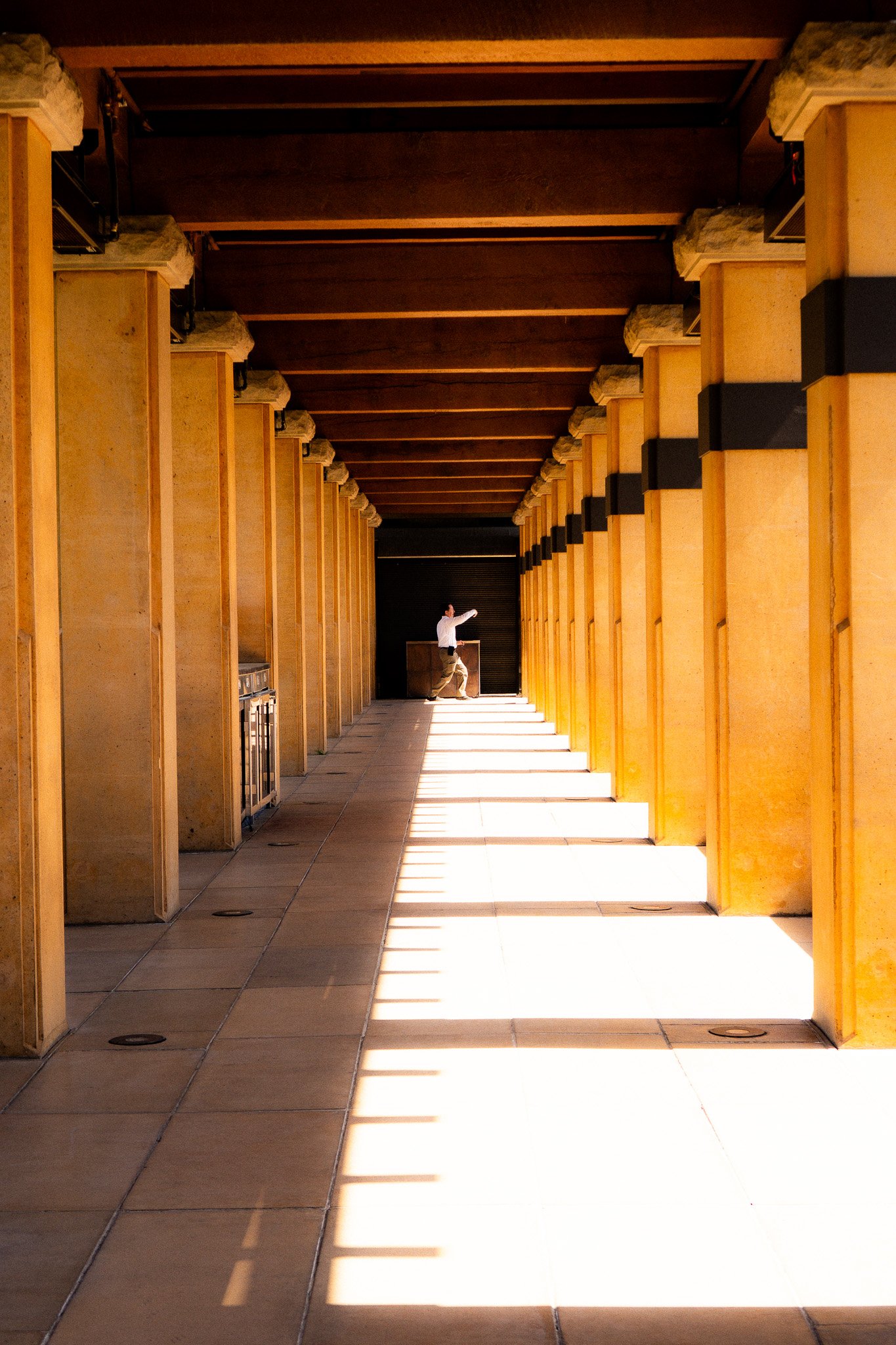 A person walking through a long corridor with tall columns on either side, sunlight creating striped shadows on the floor.