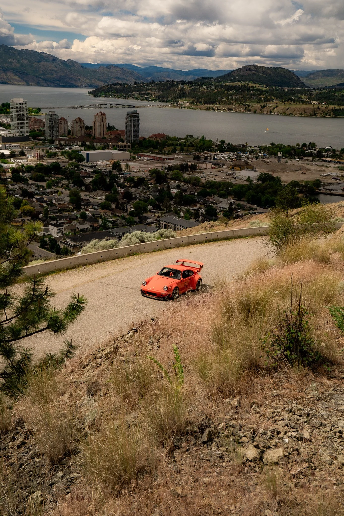 A bridge over a river with city buildings, mountains, and a cloudy sky in the background; a red sports car on a rugged dirt road in the foreground.