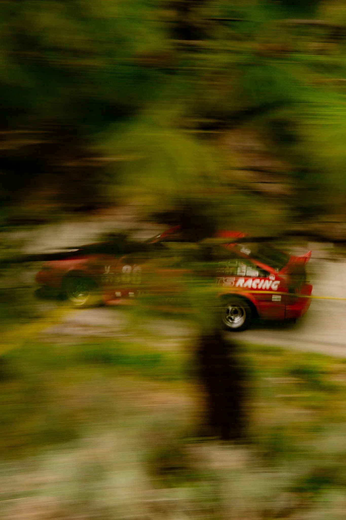 A red race car with black and white markings traveling at high speed on a racetrack, creating a motion blur effect.