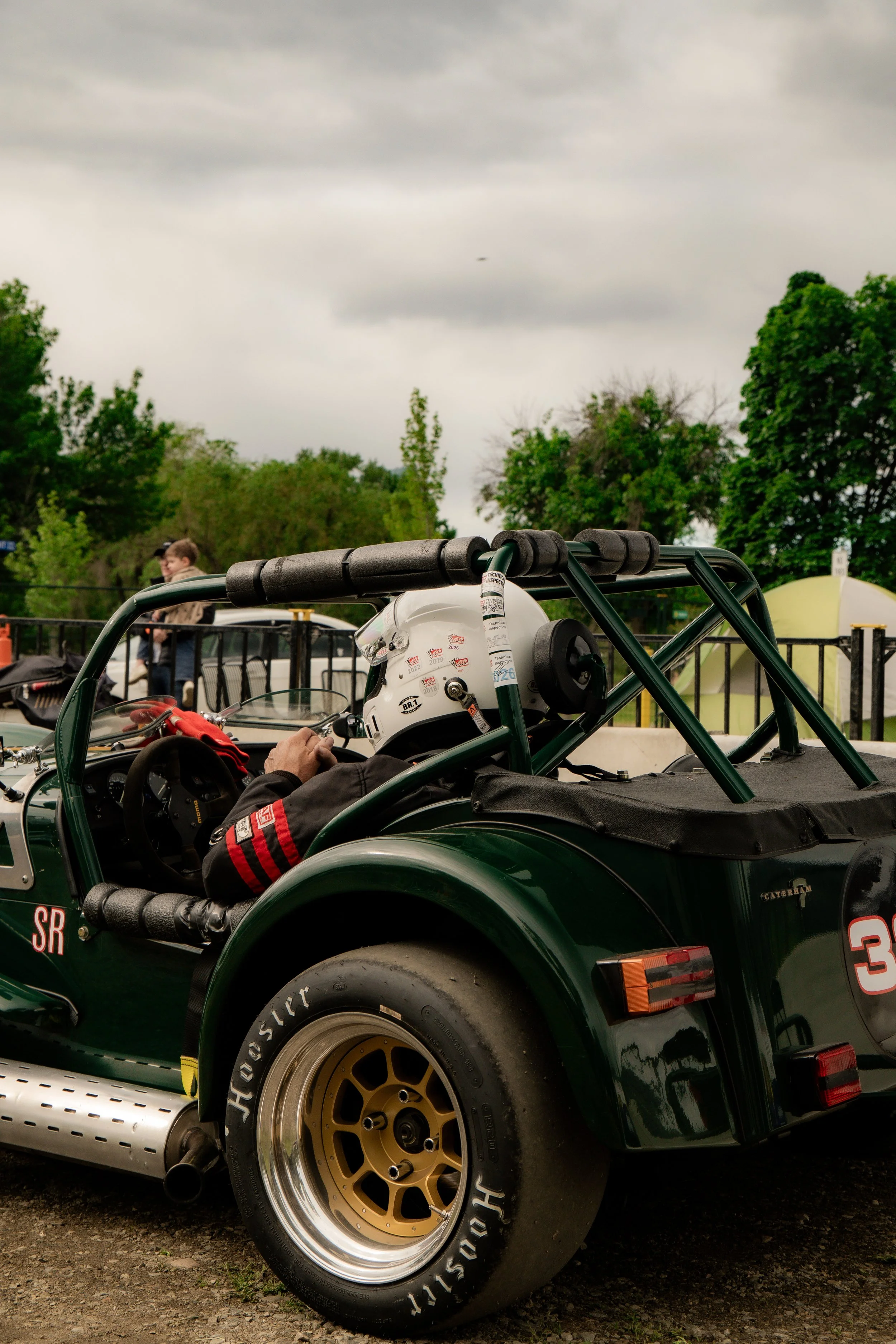 Close-up of a green race car with gold wheels and a white helmet on the driver's seat, set outdoors on cloudy day with trees and people in the background.