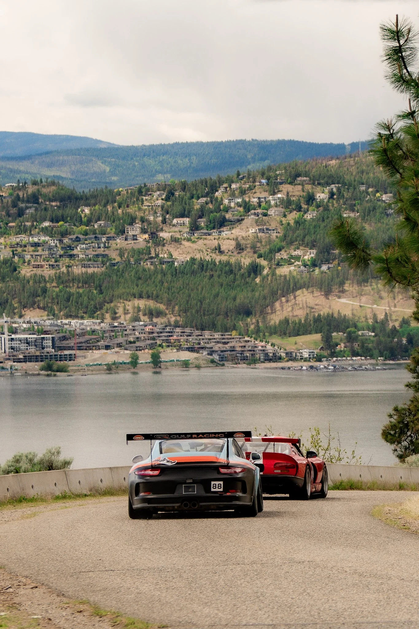 Two sports cars, a black Porsche and a red Ferrari, parked on a roadside overlooking a river and a hillside with houses and trees, under a cloudy sky.