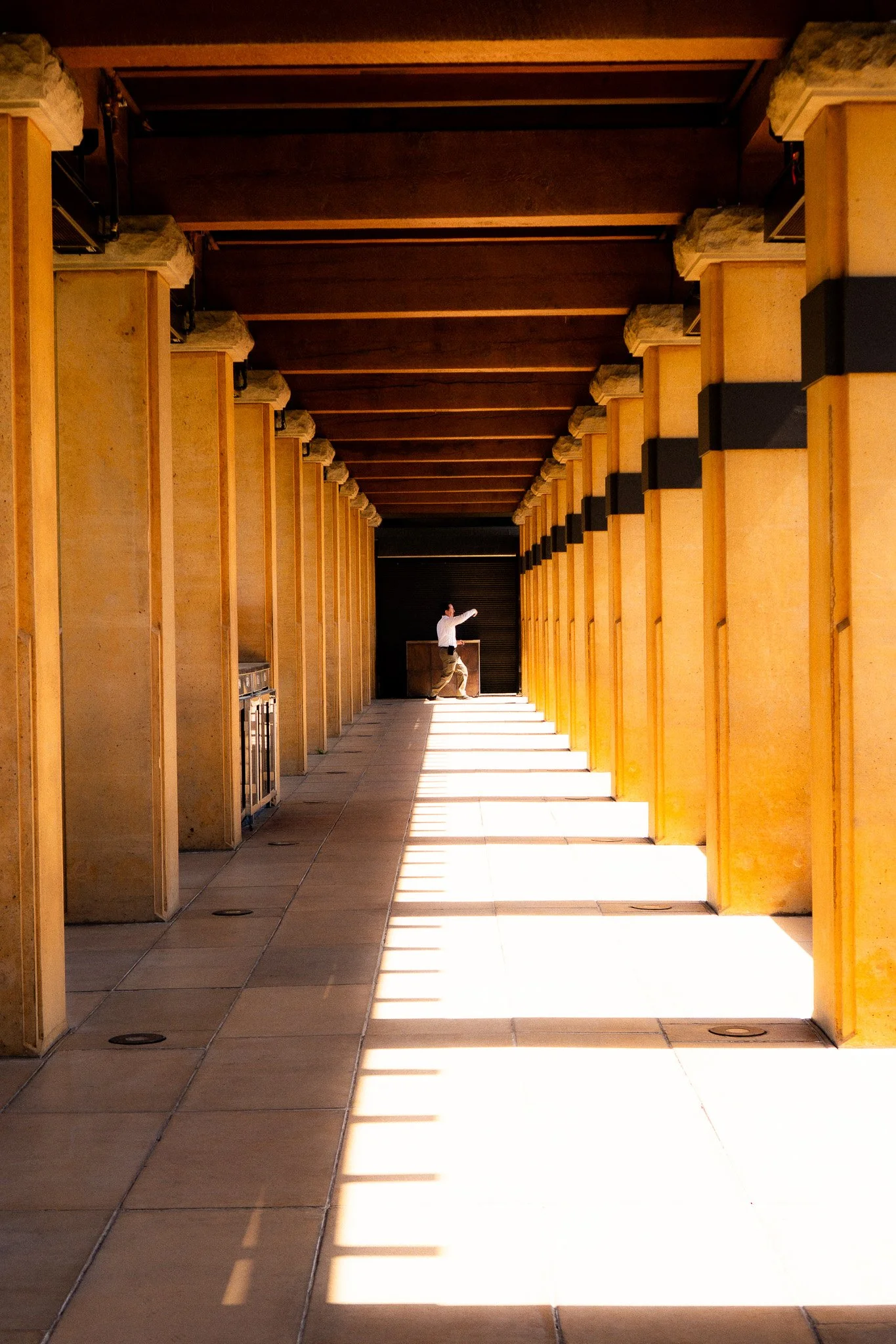 A person in a white shirt stands at the end of a sunlit corridor with stone columns. The light creates a pattern of shadows on the floor.